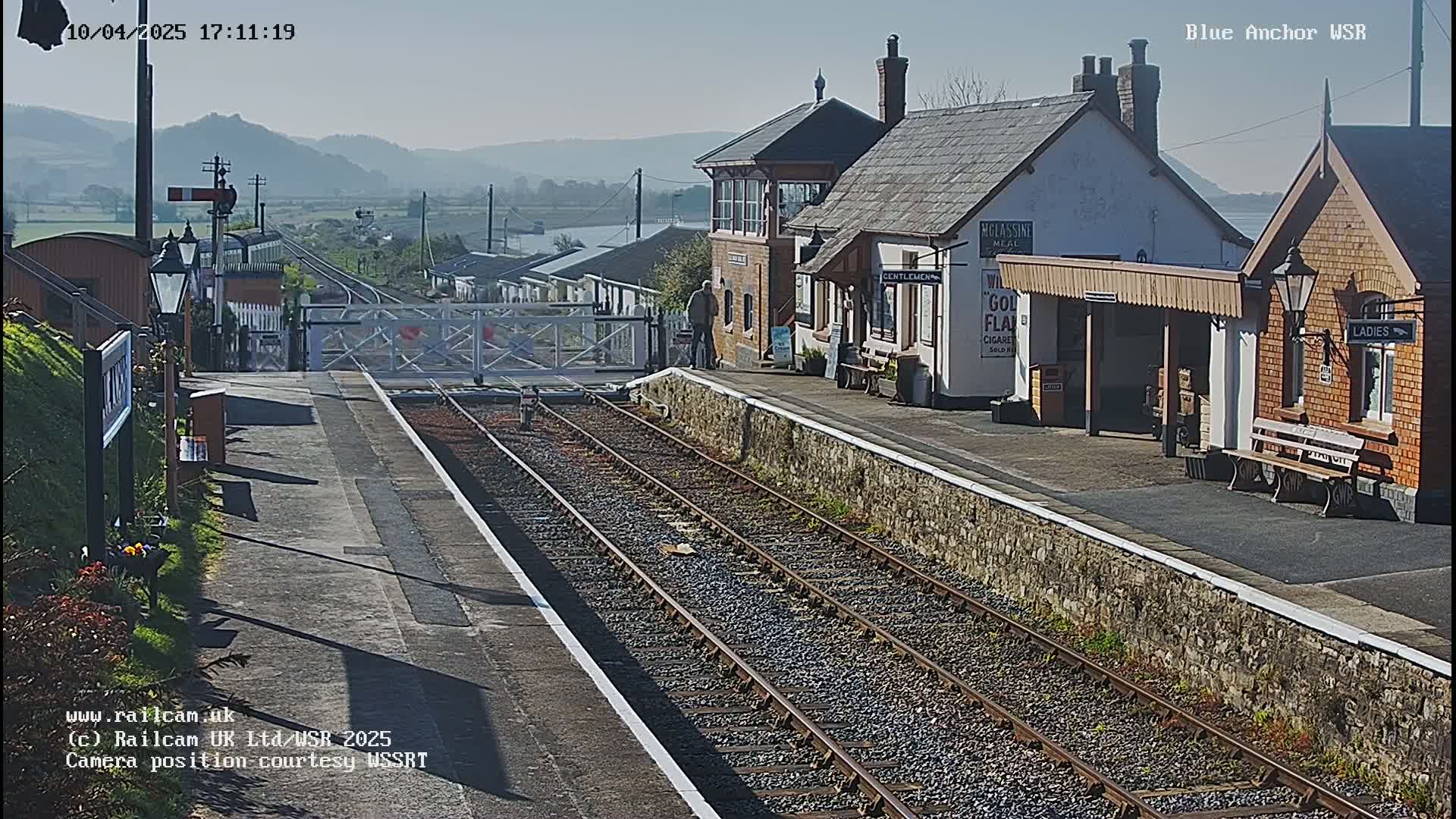 A sunny view of a train station with a signal box and passenger shelters, set against a backdrop of distant hills.