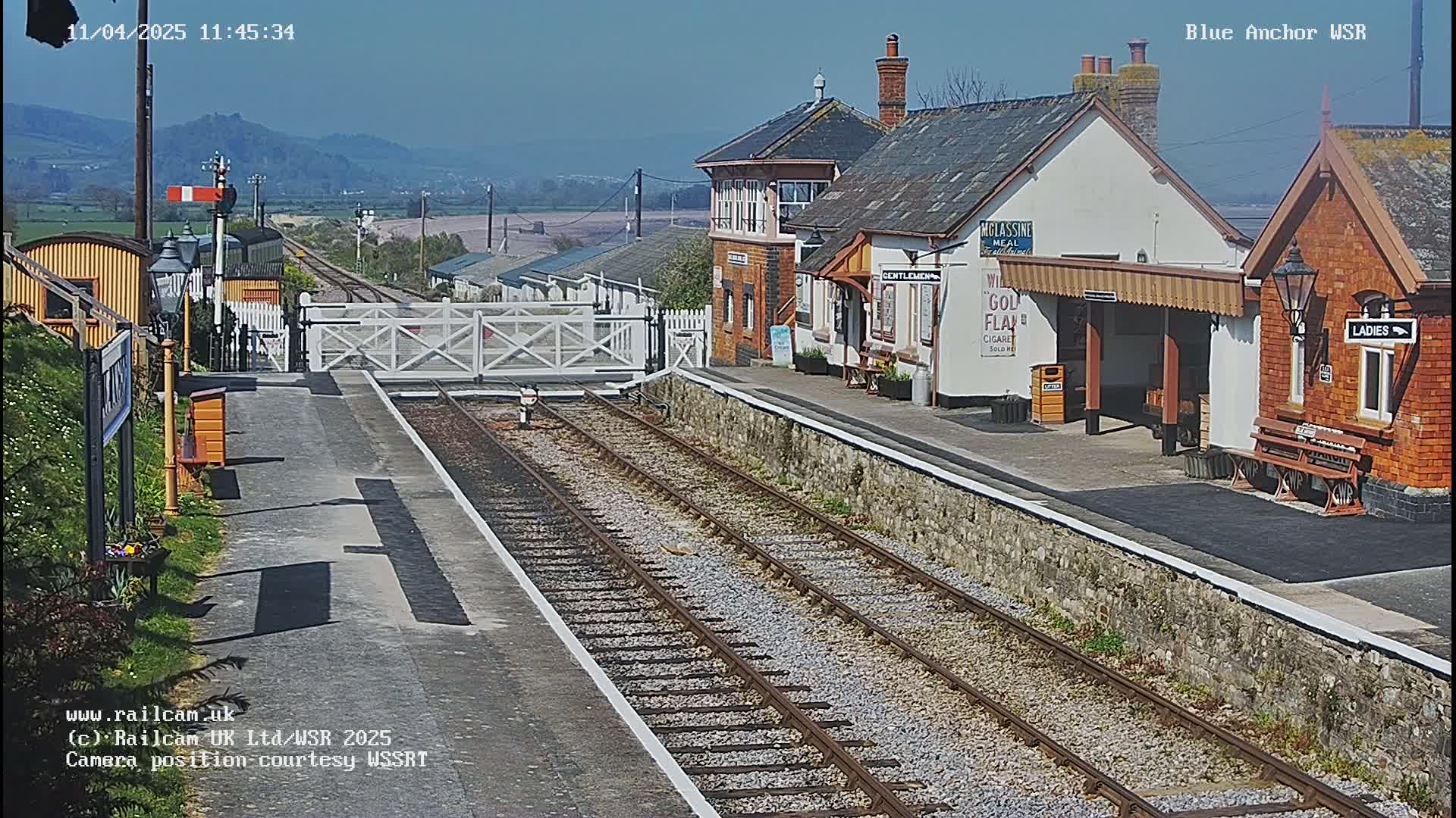 A sunny view of a train station with two tracks, a signal, and several small buildings, including a waiting area.