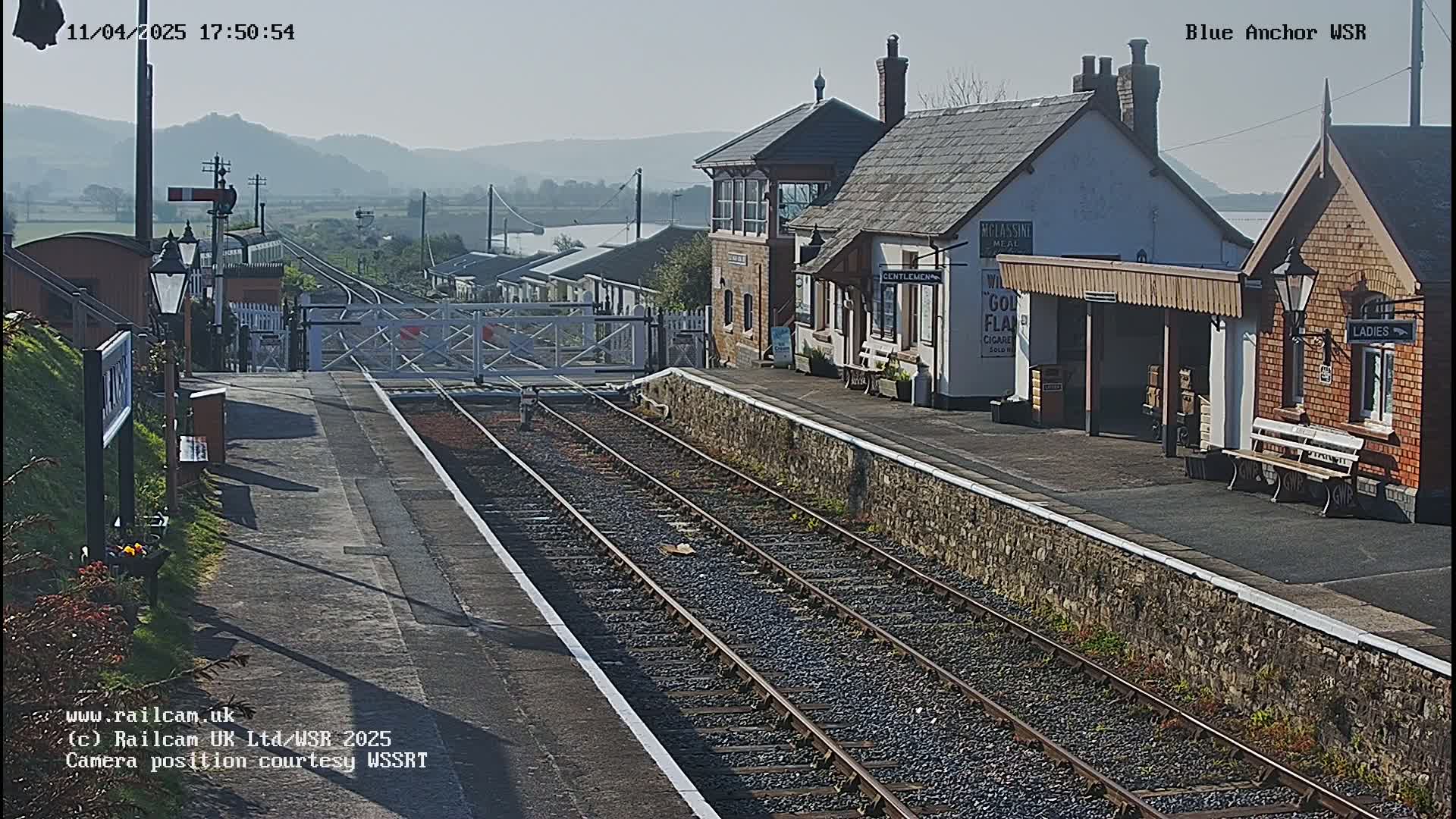 A sunny view of a train station with two parallel train tracks leading to a closed level crossing, and buildings including a signal box and waiting shelters.