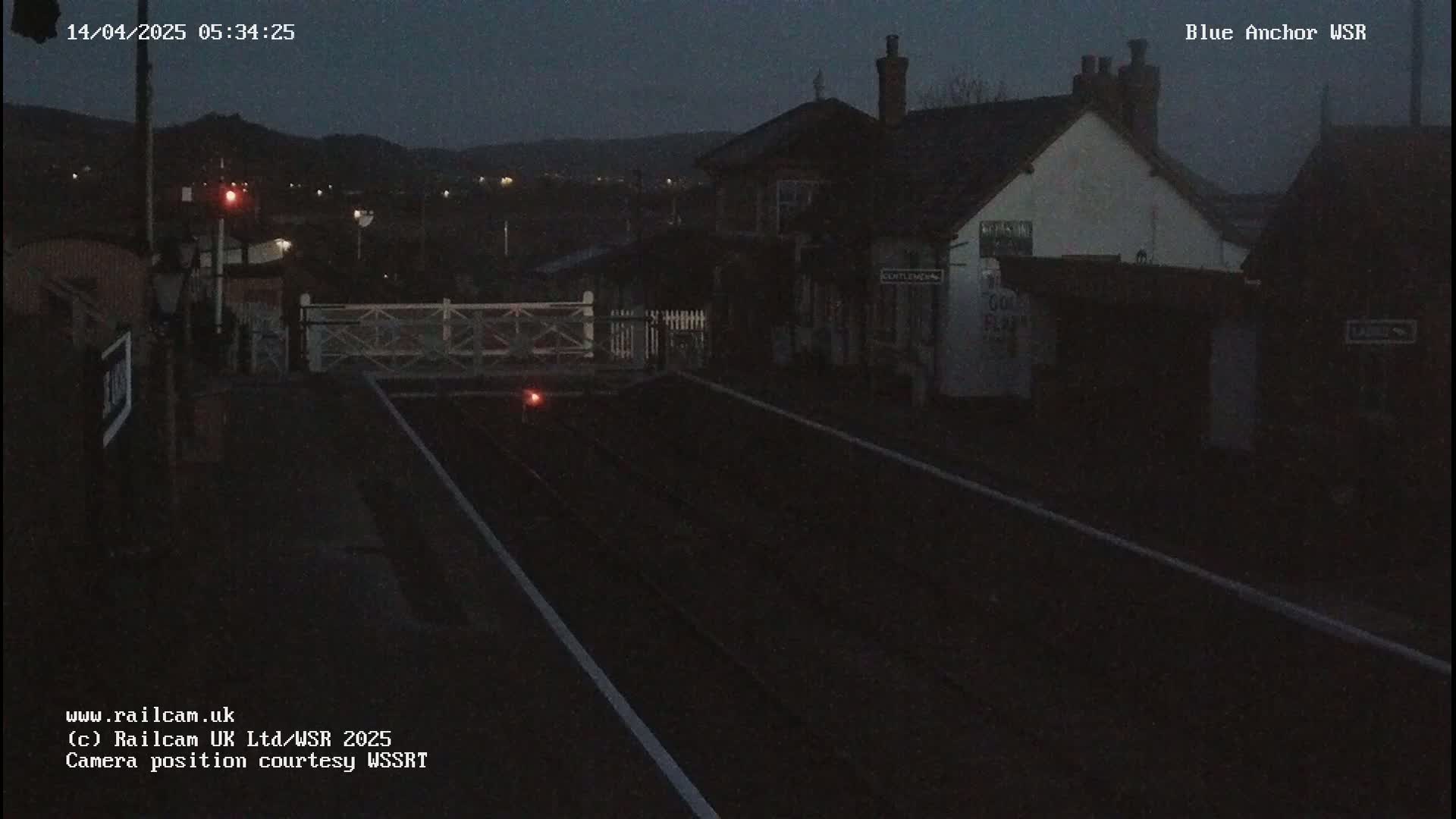 A dark, nighttime view of a train station with a closed crossing gate, under a mostly clear sky.