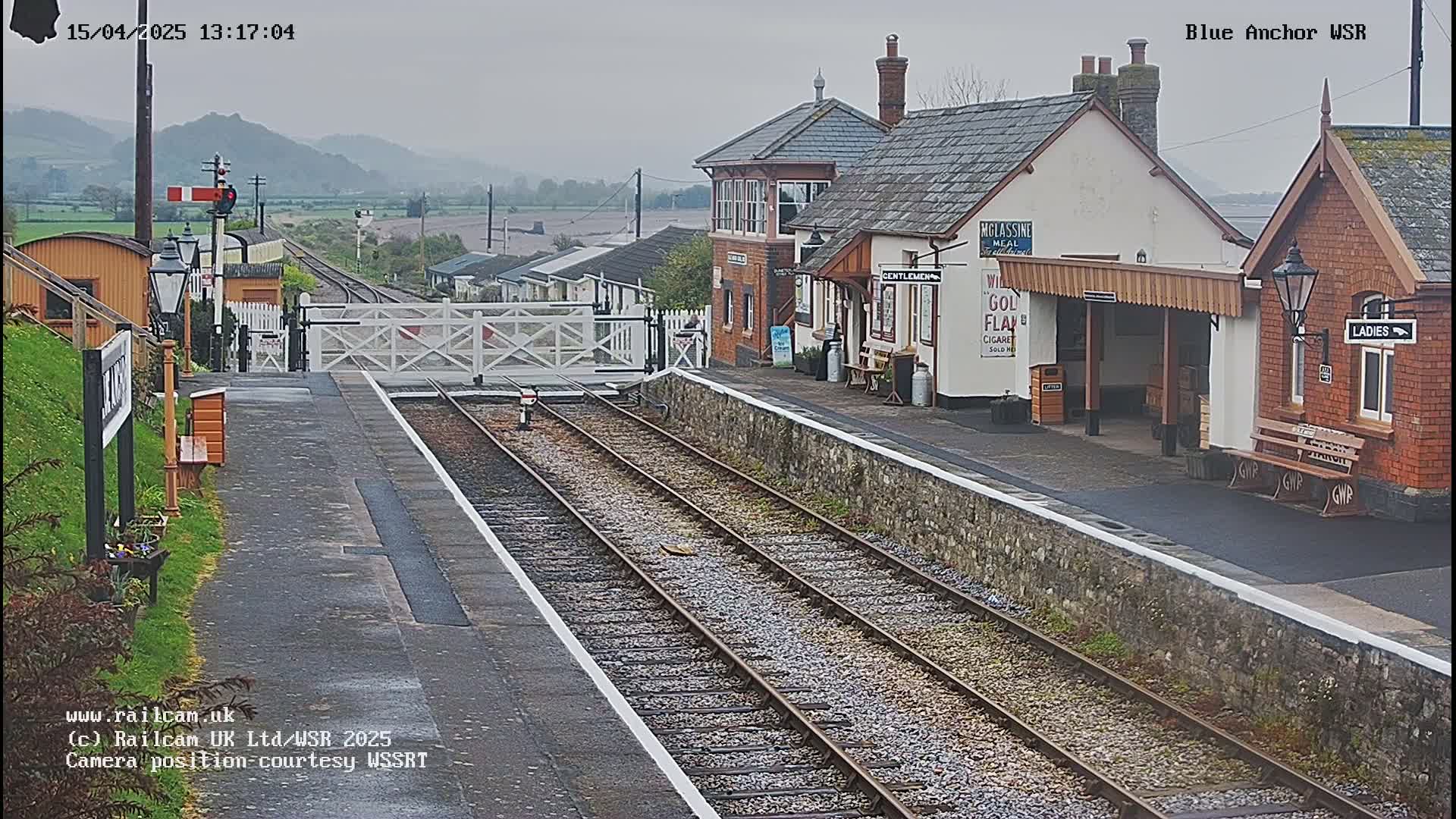 A railway station with several buildings, a closed level crossing, and two train tracks, on an overcast day.