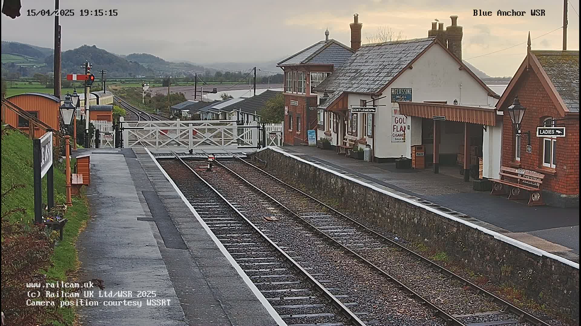 A train station with a train visible in the distance, under an overcast sky, is seen from the perspective of the tracks.