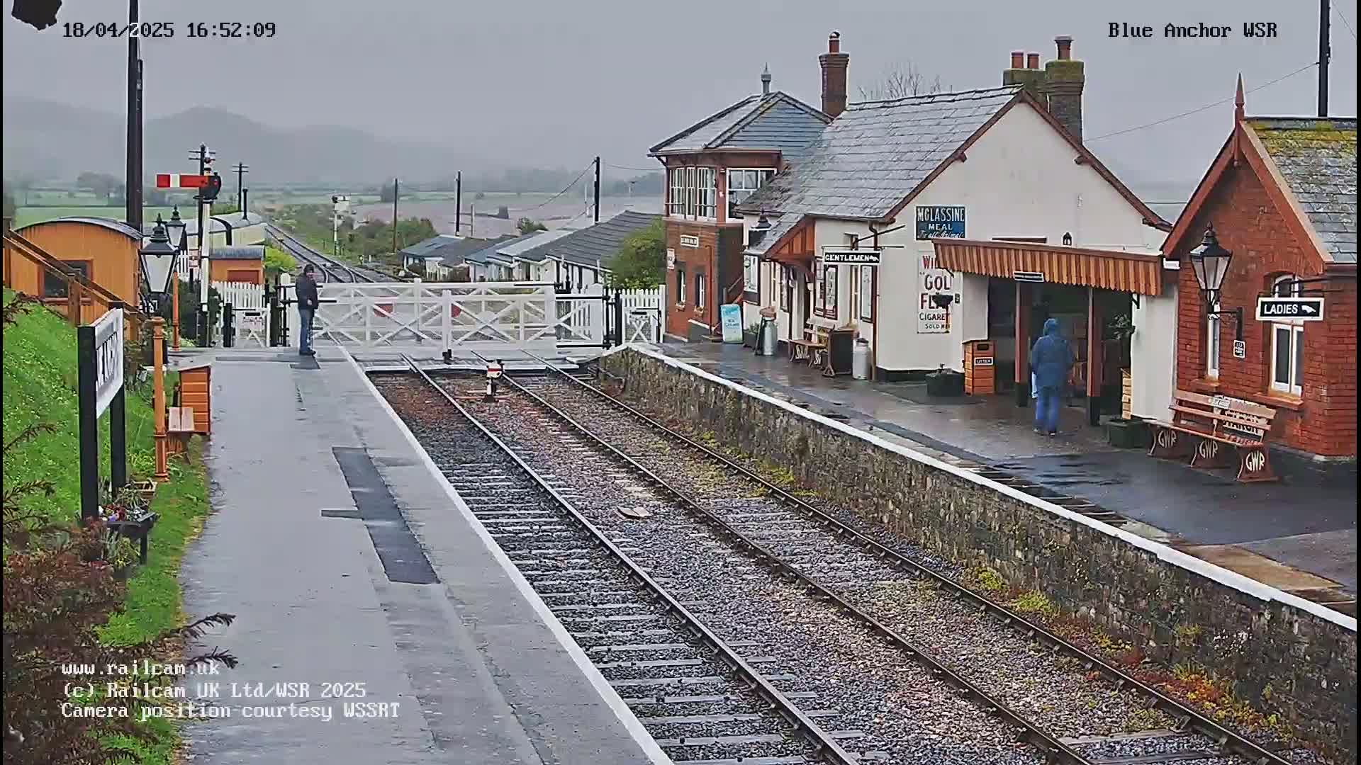 A wet, overcast day at a train station with two railway tracks leading to a small building and a waiting area.