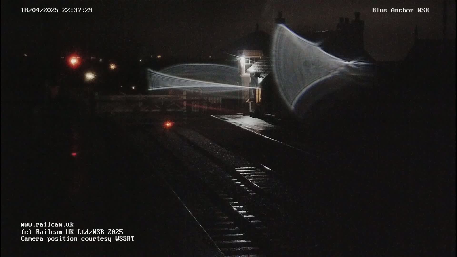 A nighttime view of a train station with snow on the ground, showing train tracks and a building illuminated by lights, with blurry streaks of light possibly indicating a moving train.