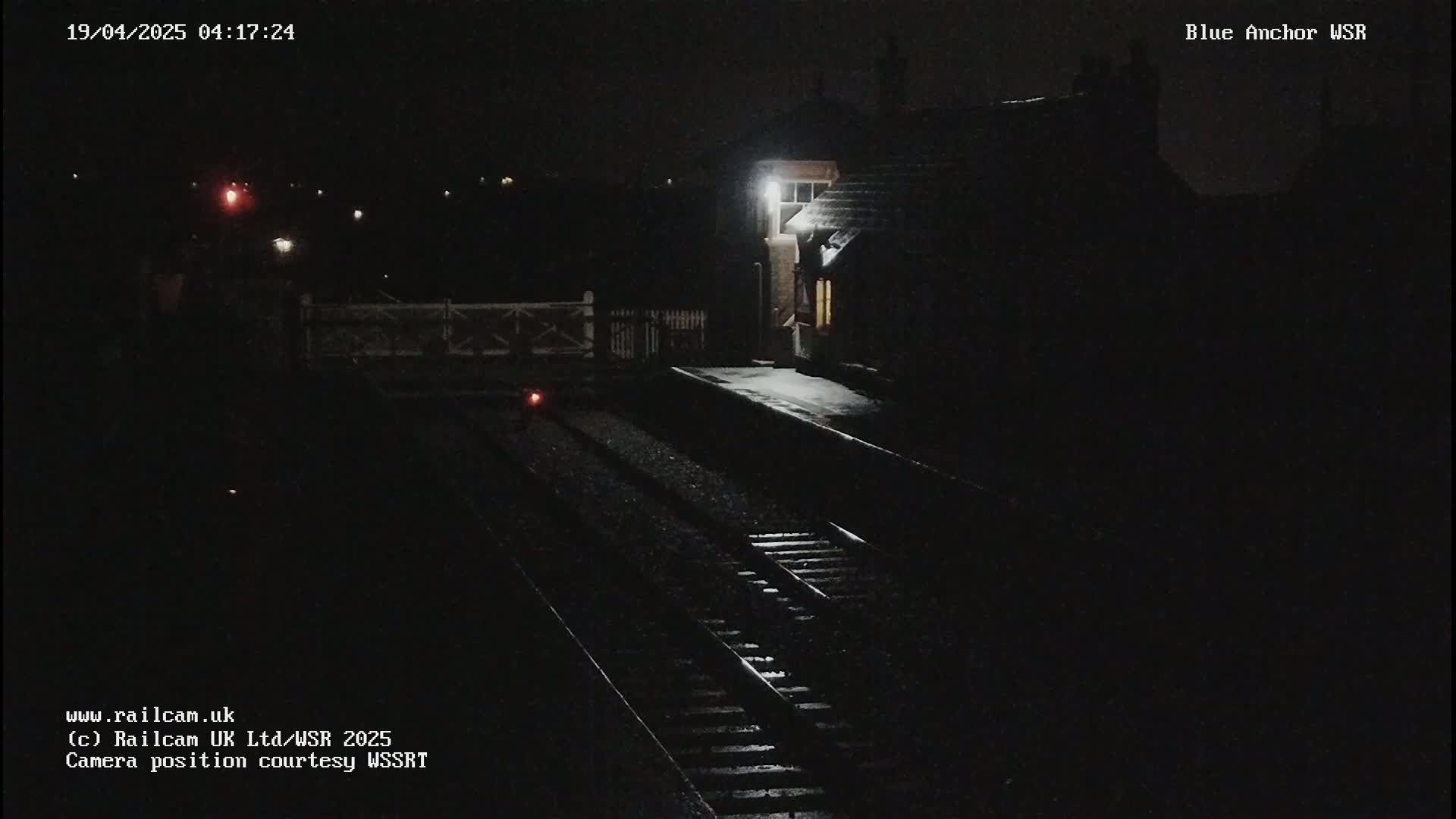 A nighttime view of a snow-covered train station platform and tracks, illuminated by a single light.