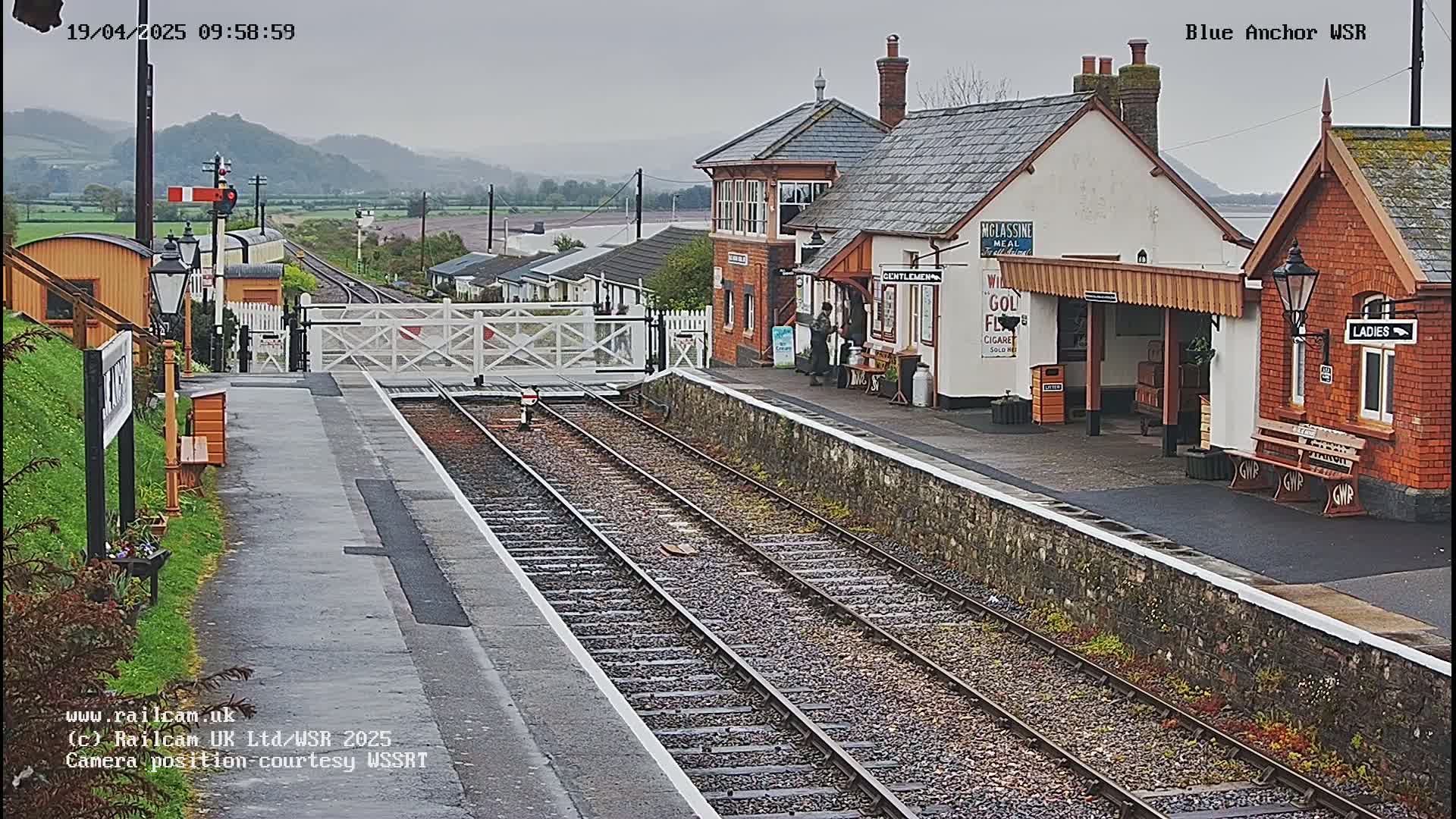 A view down a railway track at a station on an overcast day, showing a railway platform with station buildings.