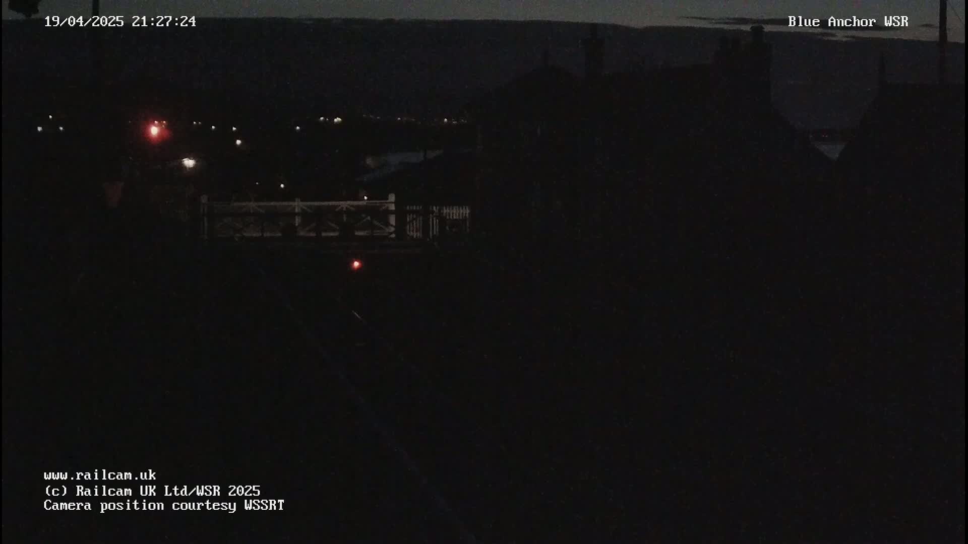 A nighttime view of a dark landscape with a few faint lights visible in the distance, and a structure resembling a fence or railing in the foreground.