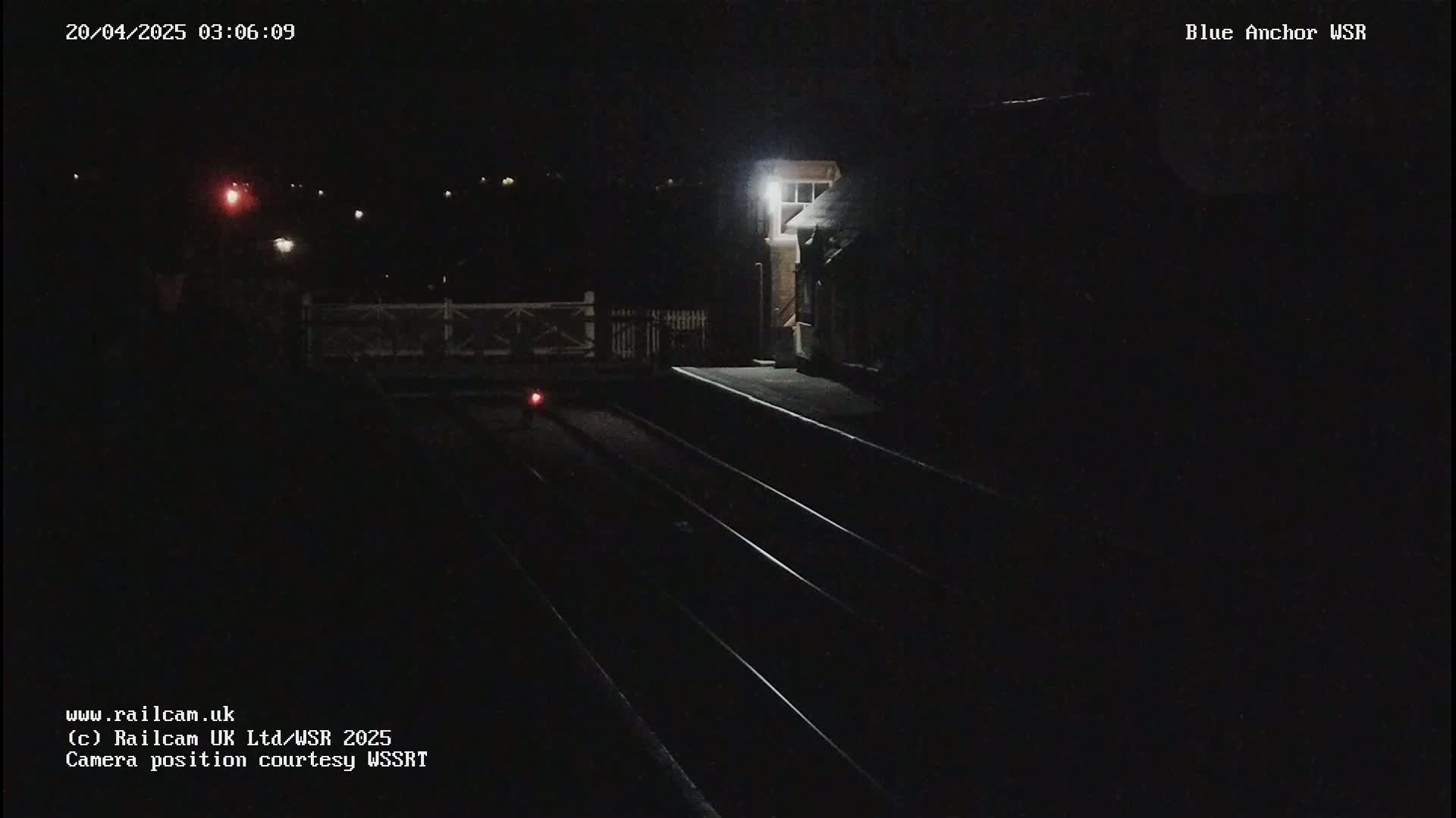 A dark nighttime view of a train station platform with train tracks and a dimly lit building.
