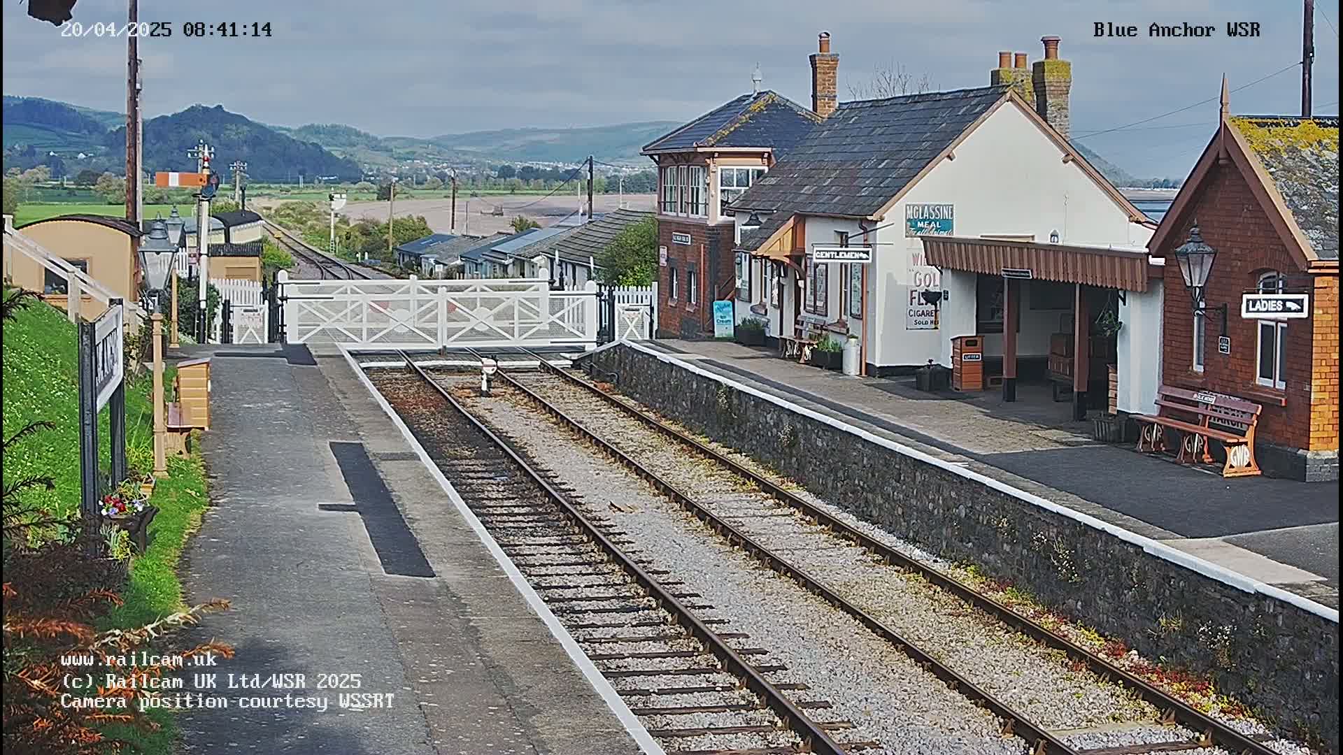 A train station with a low stone wall separating the platform from the tracks, under a mostly cloudy sky.