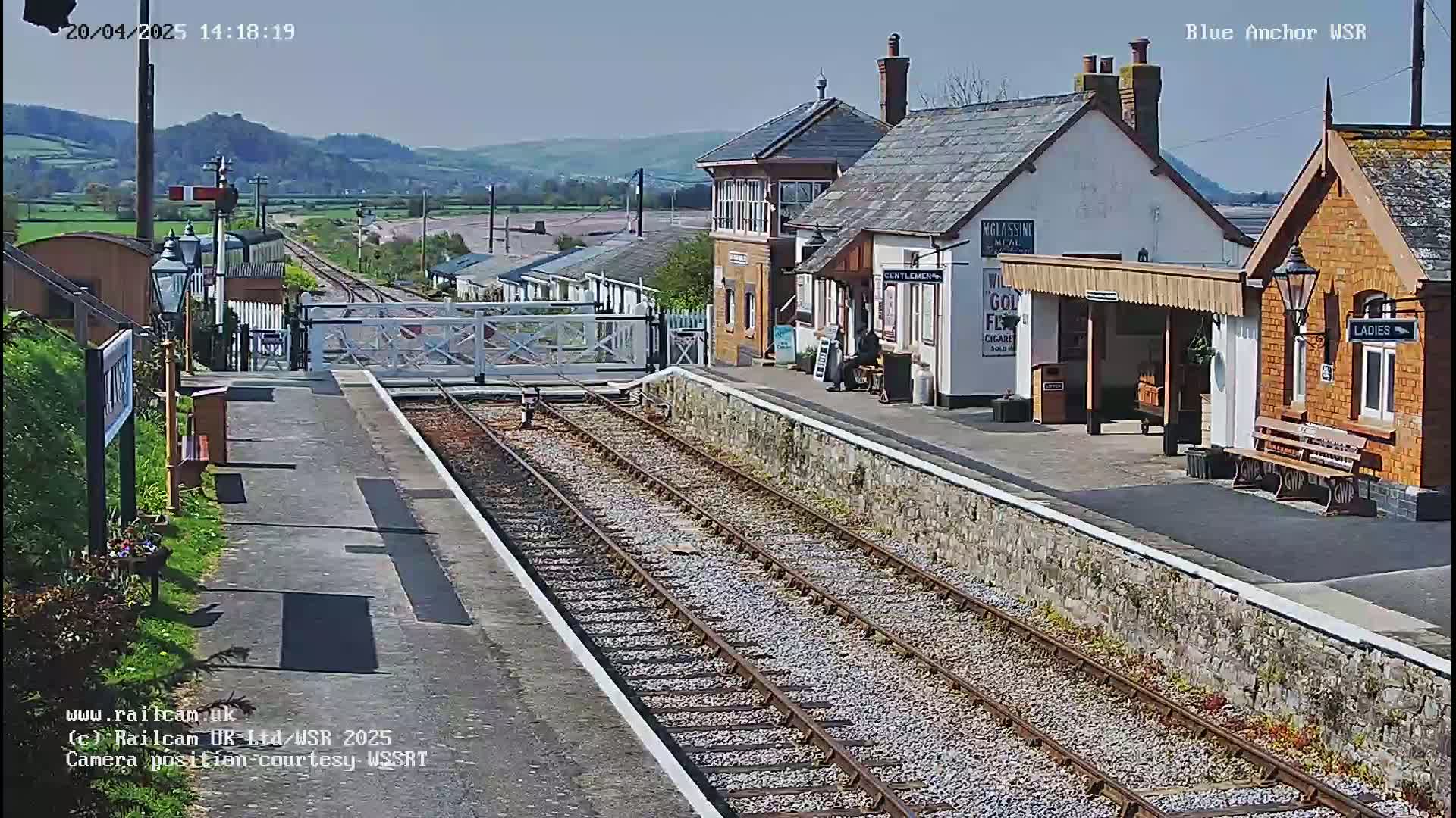A sunny view of a train station with two platform buildings, railway tracks, and a level crossing gate, set against a backdrop of rolling hills.