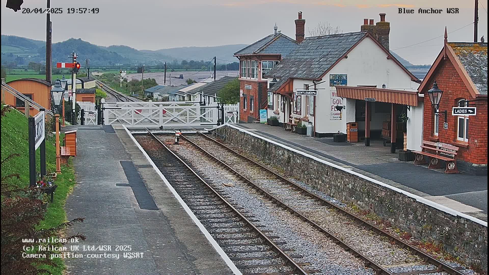 A railway station with two passenger shelters and train tracks sits under an overcast sky, with hills visible in the background.
