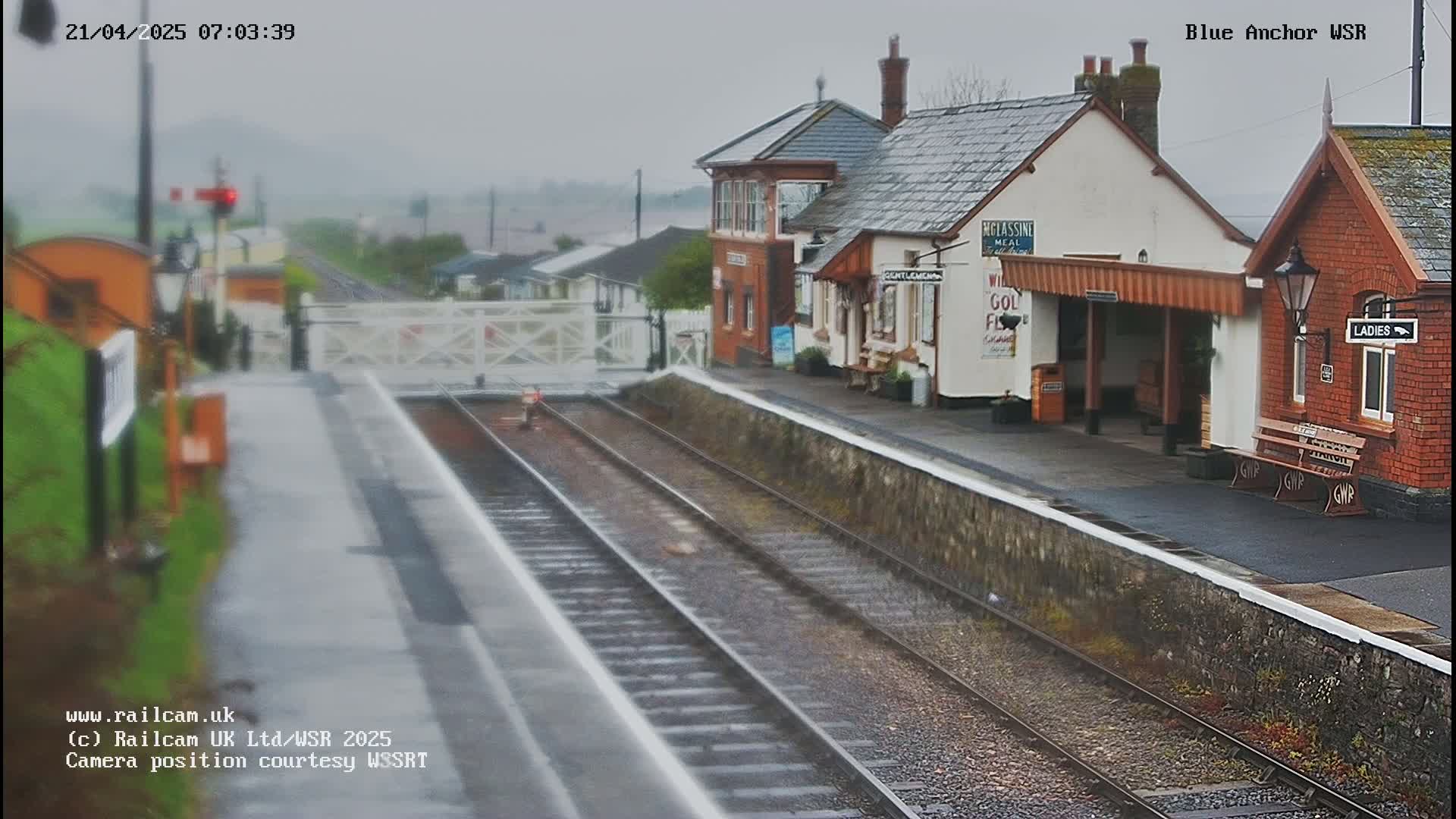 A rain-soaked railway station with two buildings and a waiting area, viewed from the tracks.