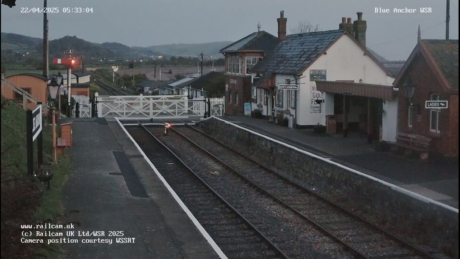 A train station with two sets of tracks and station buildings is shown under a dusky sky.