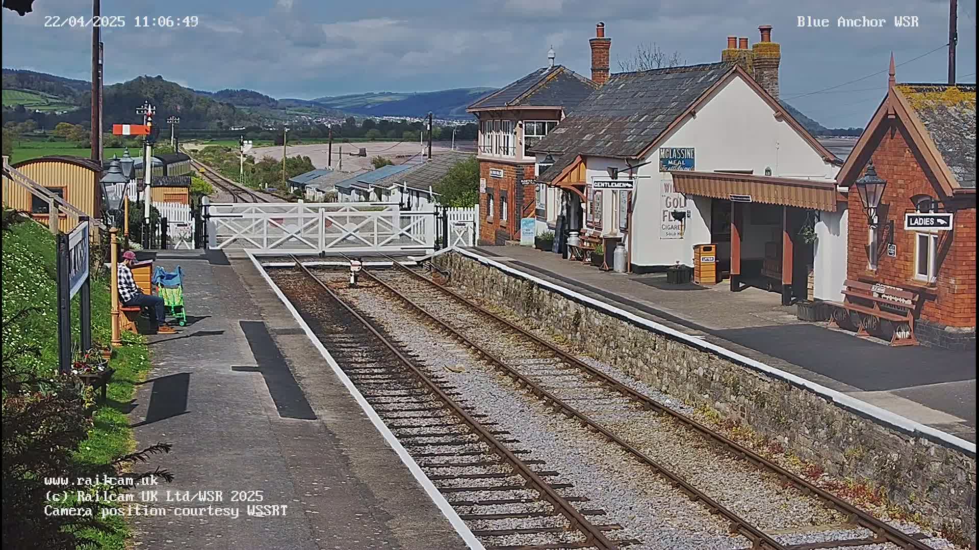 A sunny day at a train station shows a person sitting on a bench near railway tracks that lead to a small collection of buildings.