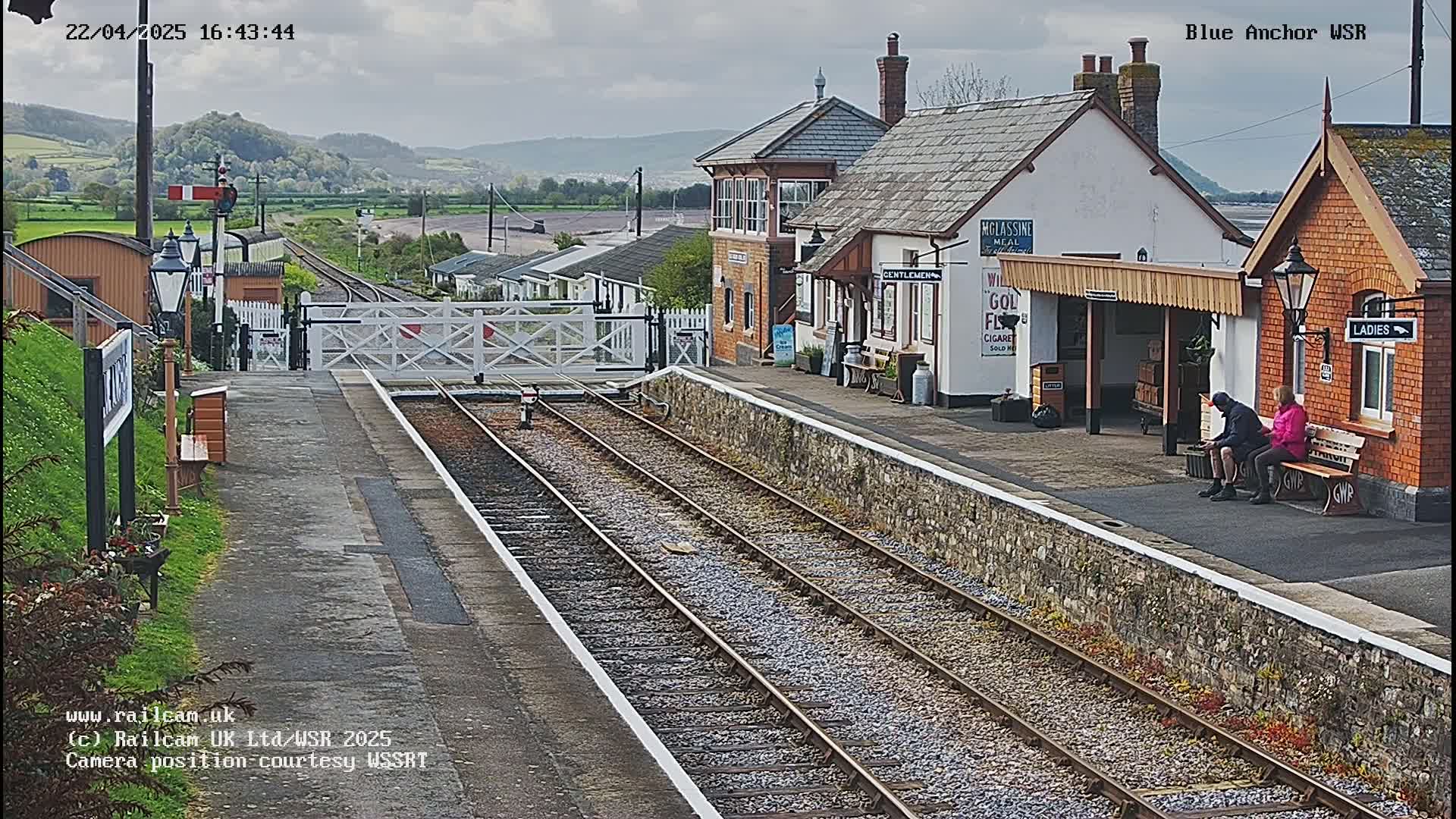 A railway station with two people sitting on a bench, under a partly cloudy sky, features a stone wall separating the platform from the tracks.