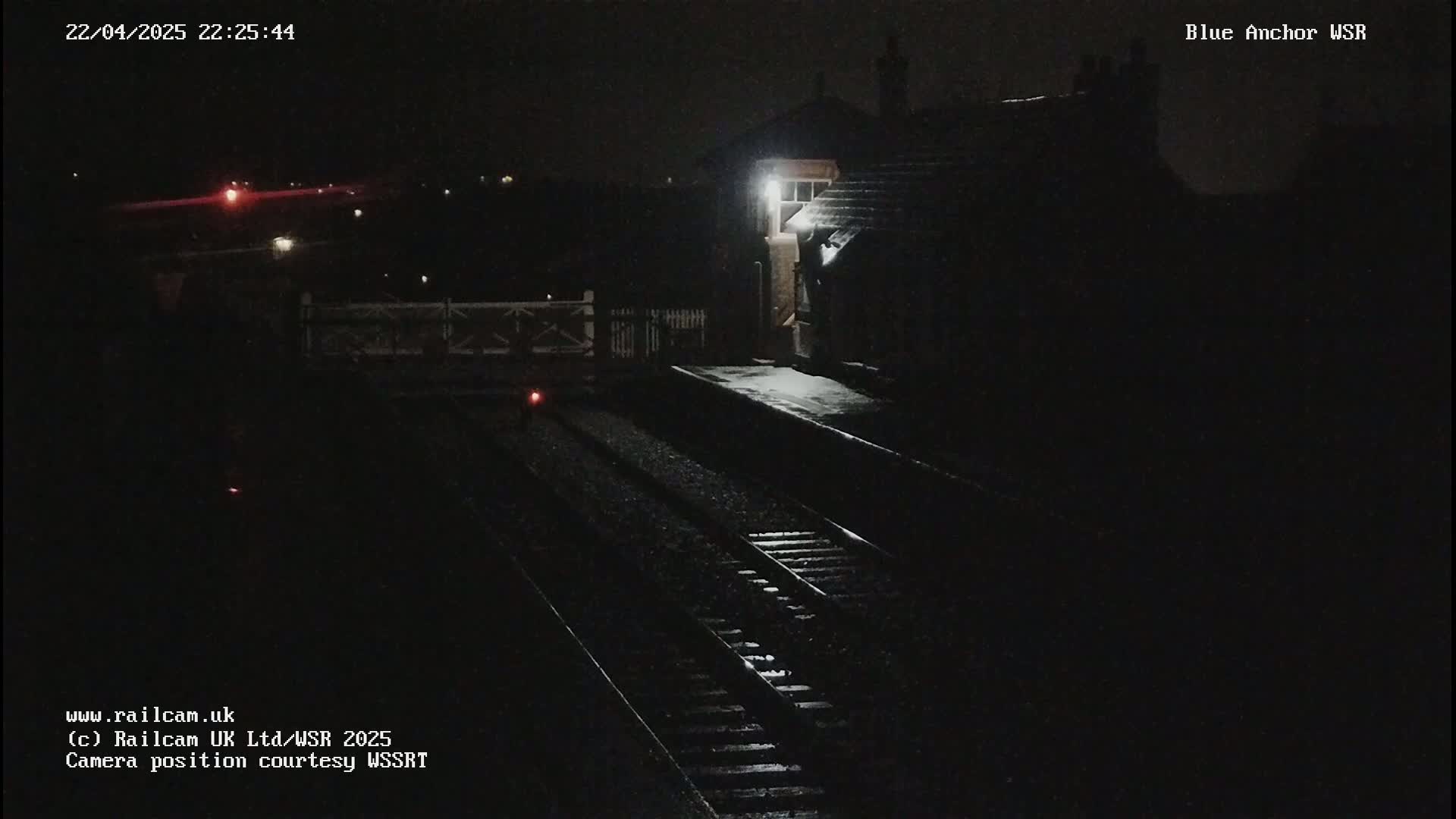 A nighttime view of a dimly lit train station platform with train tracks and a building under a dark sky.