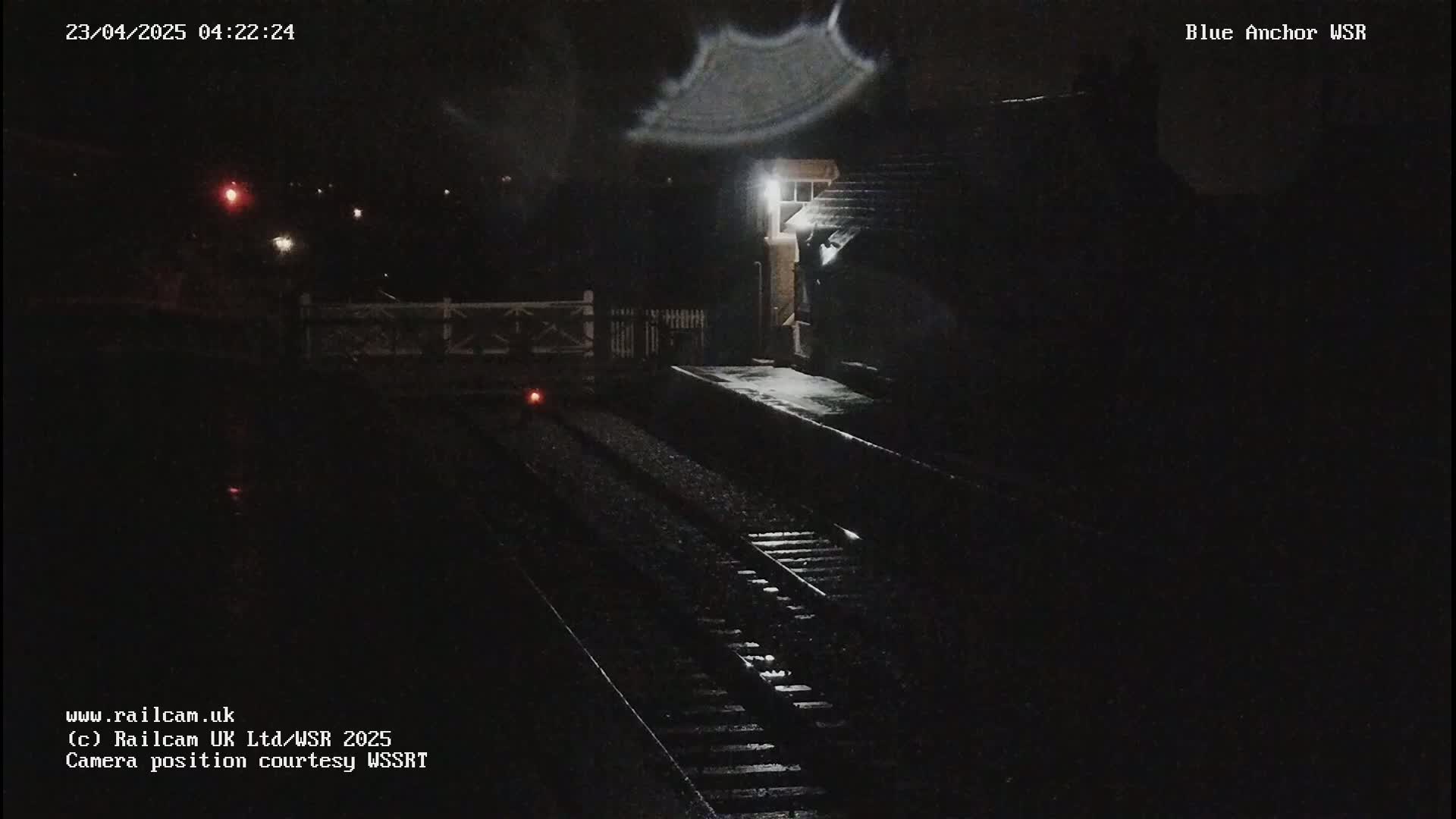 A nighttime view of a snow-covered train platform and tracks, dimly lit by a single light.