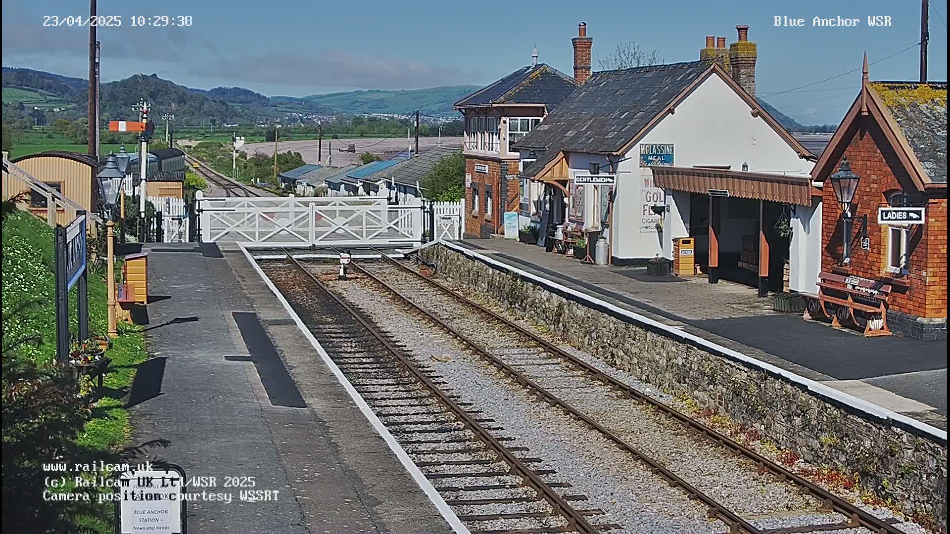 A sunny view of a train station with two sets of tracks and several buildings, including a signal box and waiting shelters.