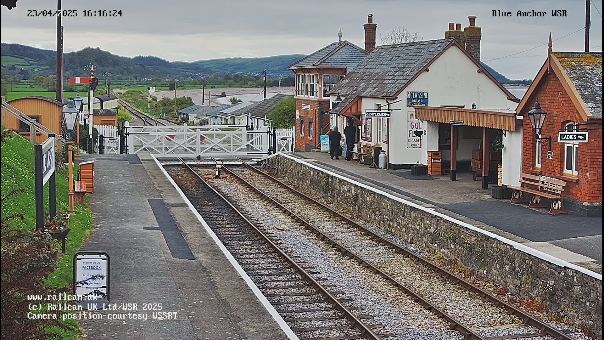 A train station with a stone platform, railway tracks, and several buildings under an overcast sky.