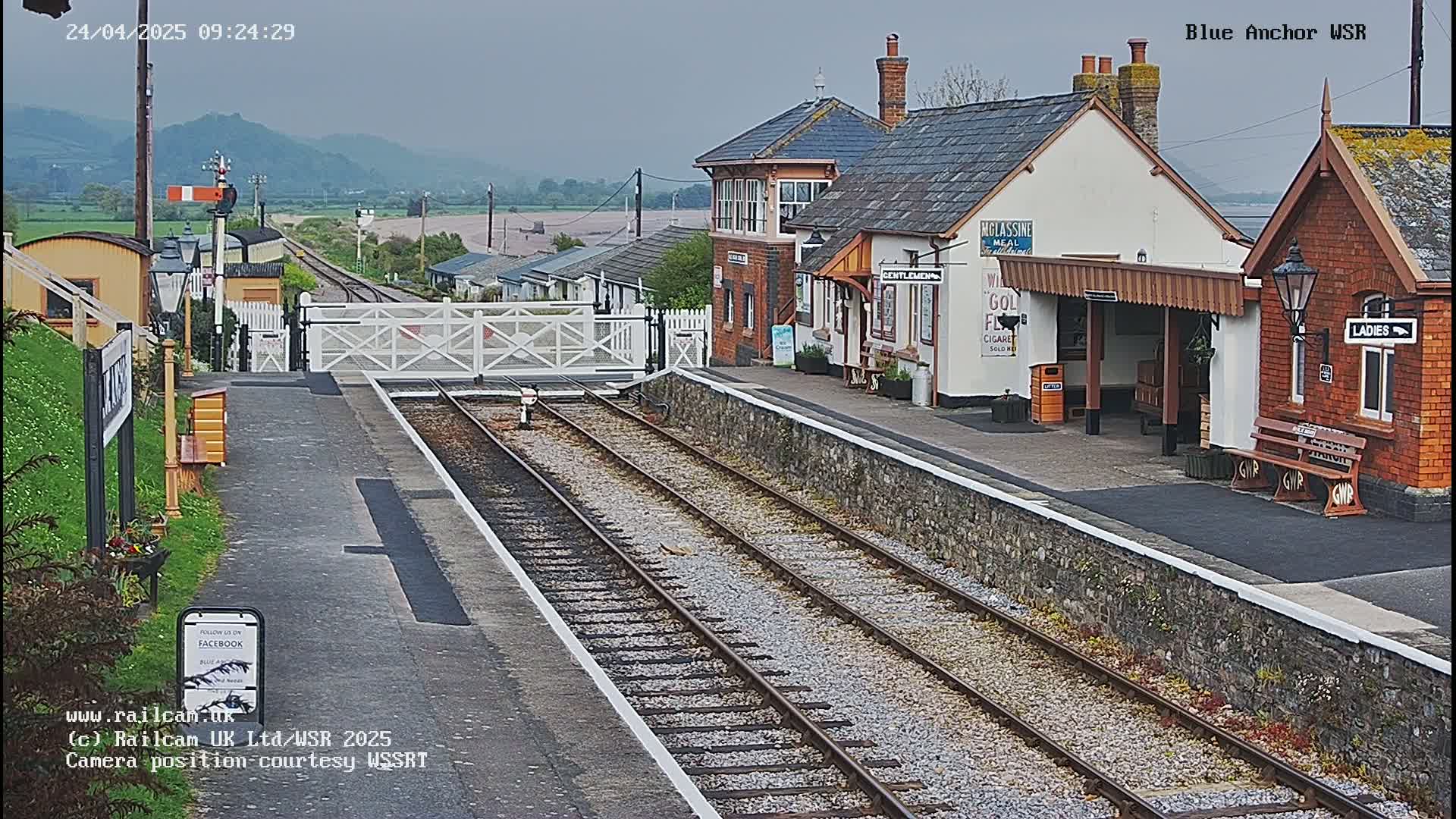 A train station with a covered platform and several buildings sits on a railway line under an overcast sky.