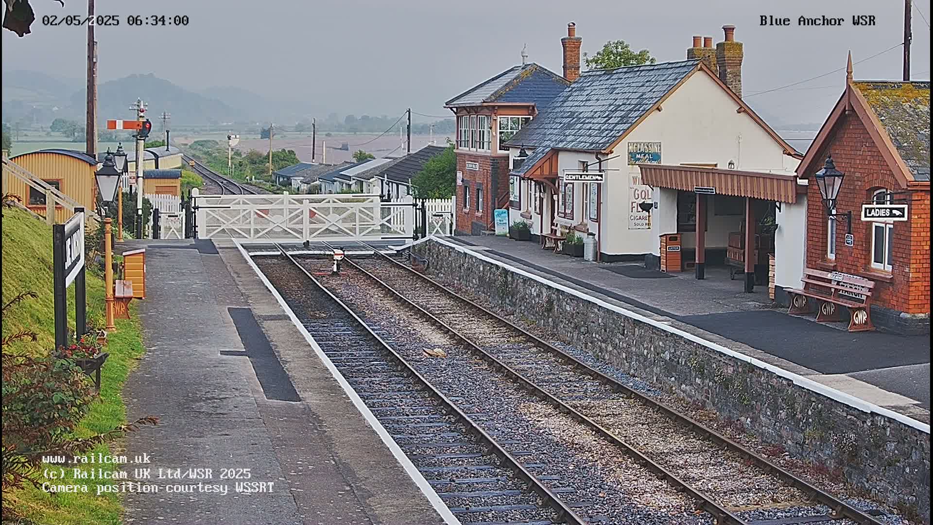 A train station with a low brick building and covered platform is seen on an overcast day, with railroad tracks extending into the distance.