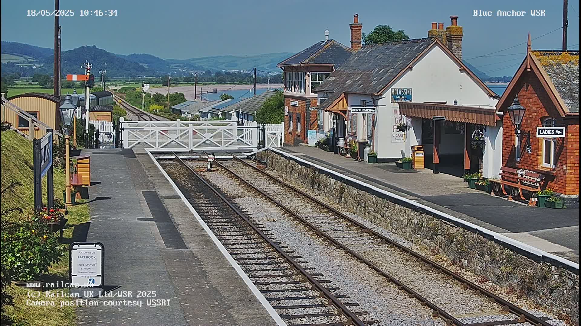 A train station with a platform, tracks, and station buildings sits on a sunny day with hills and a body of water visible in the distance.