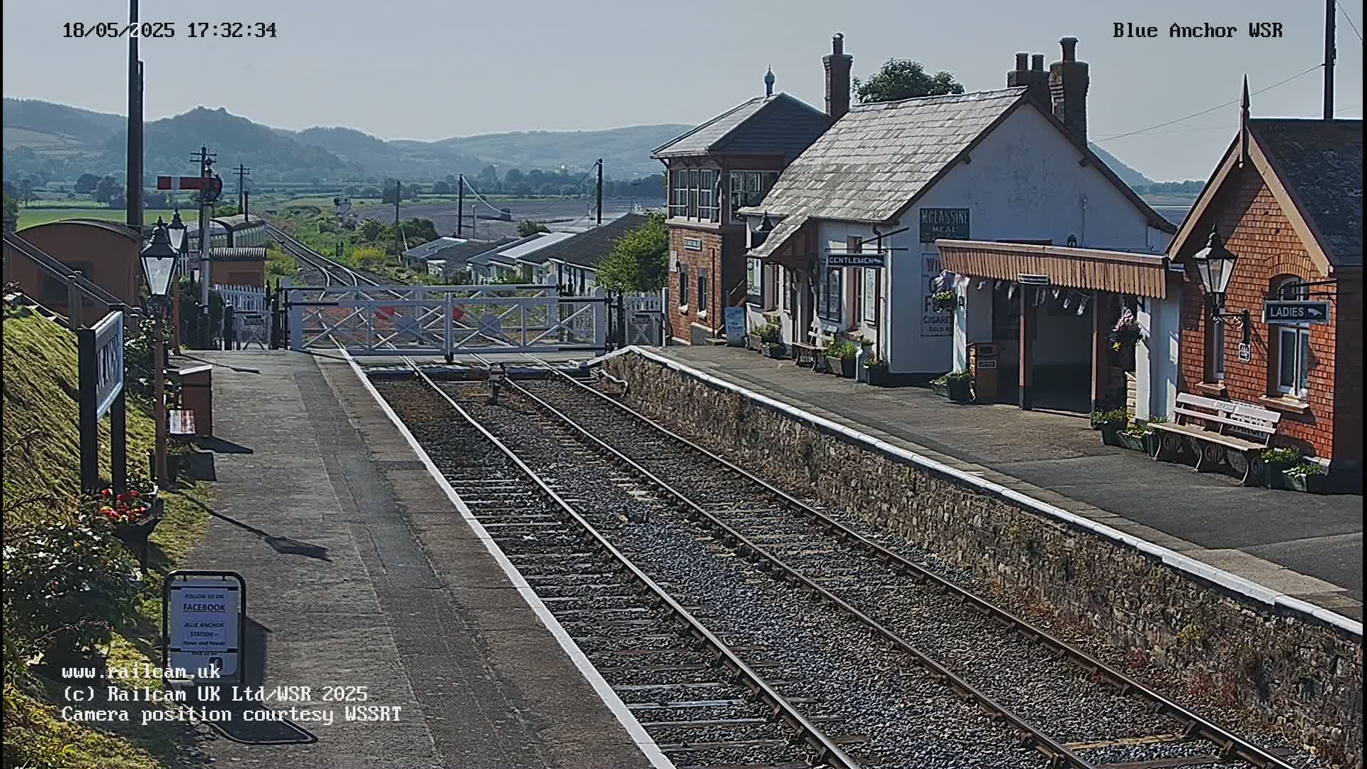 A sunny view of a train station with two sets of tracks, a signal box, and passenger shelters.