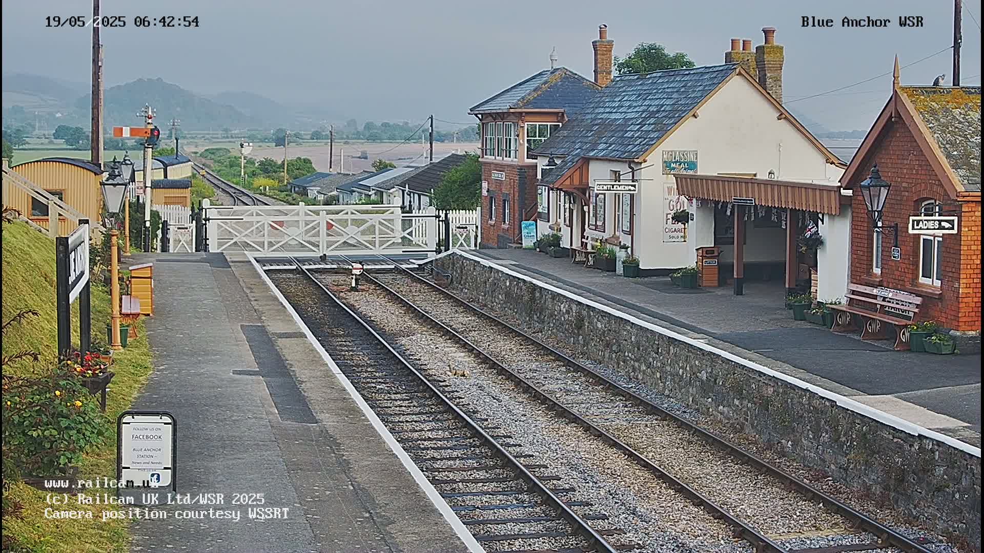 A sunny view of a train station with two sets of tracks, a signal box, and passenger shelters.