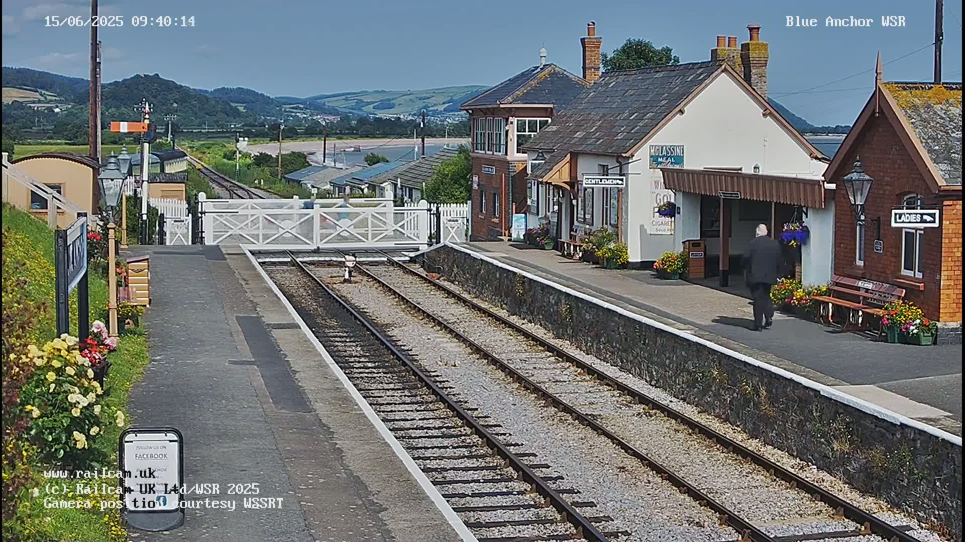 A train station with a brick building and flower boxes, situated near a body of water and hills under a clear, sunny sky.