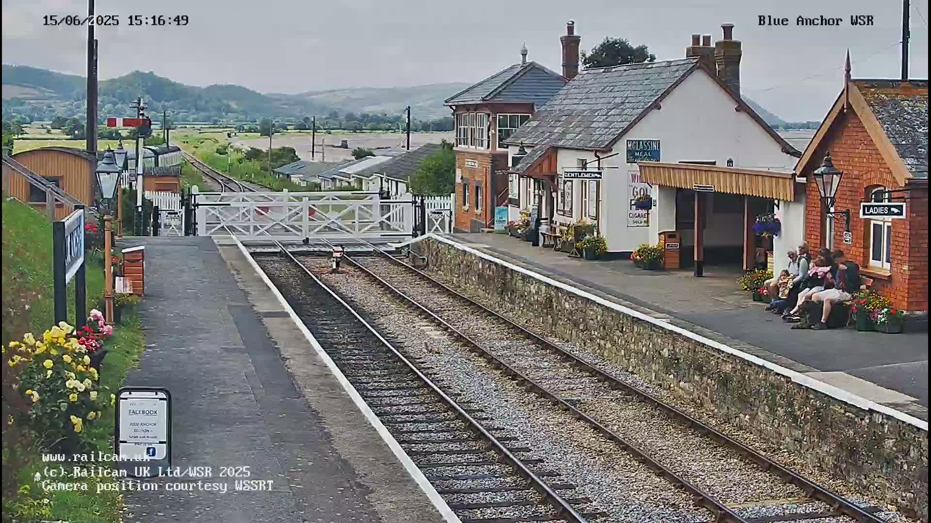 A train station with several people sitting on a bench, a closed crossing gate, and train tracks leading into the distance under an overcast sky.