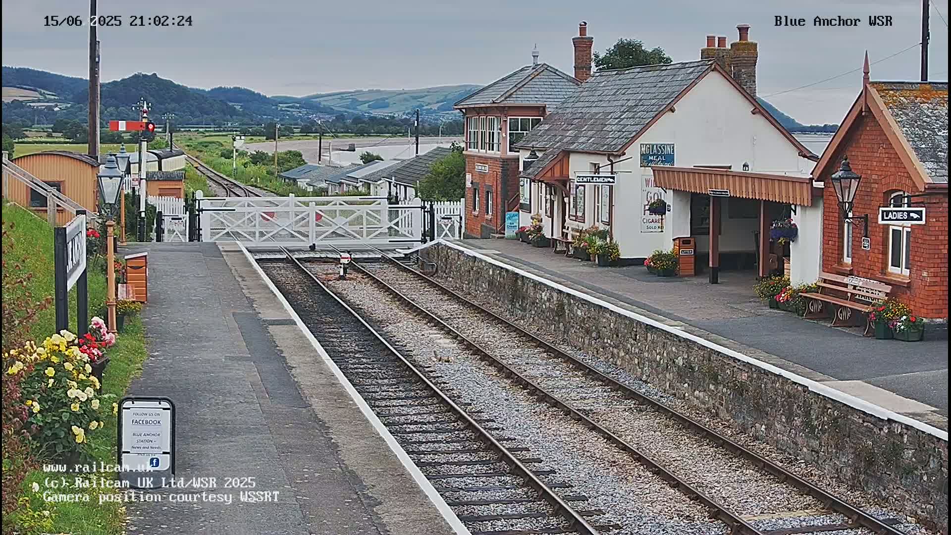 A train station with several people sitting on a bench, a closed crossing gate, and train tracks leading into the distance under an overcast sky.