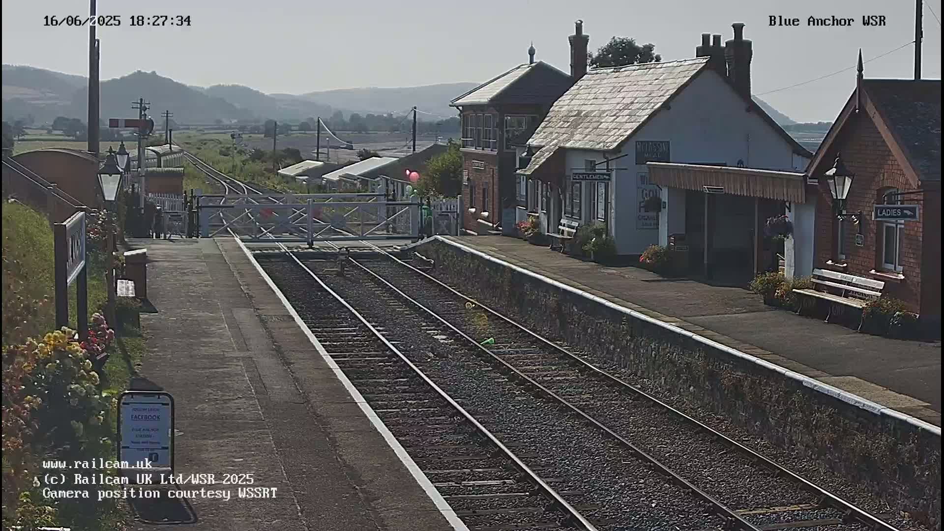 A train station with a brick building and a level crossing, under sunny conditions, with hills visible in the background.