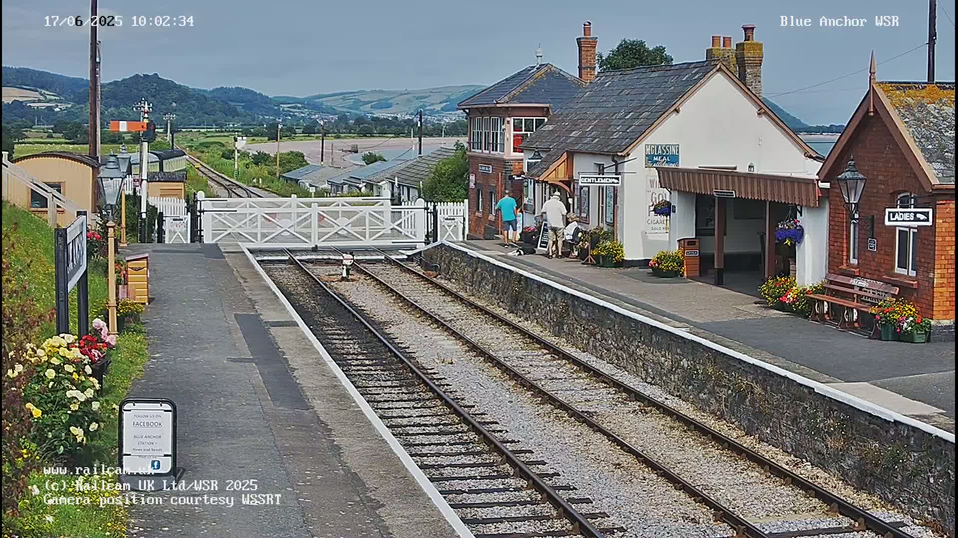A train station platform with a signal box and several buildings, under a mostly clear sky.