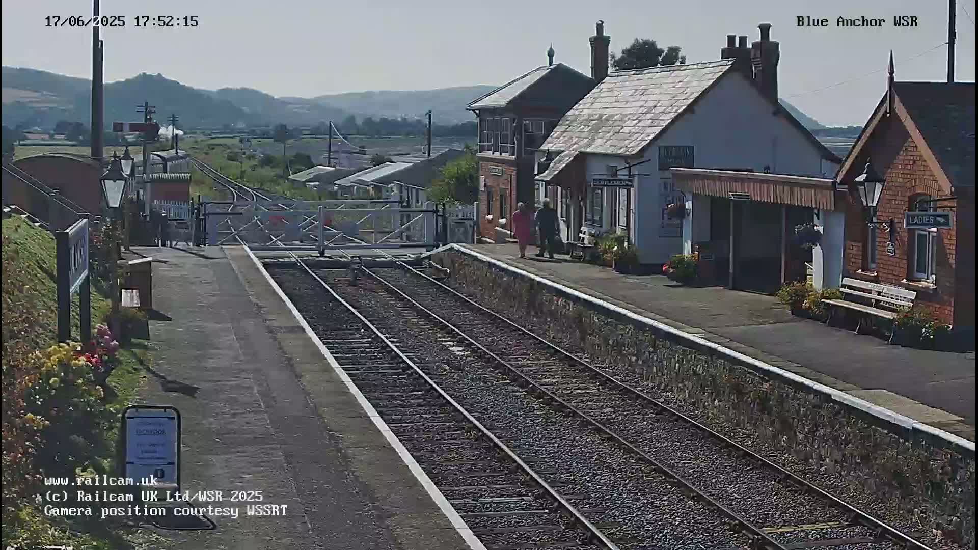 A train approaches a small, quaint railway station on a sunny day, with rolling hills visible in the background.