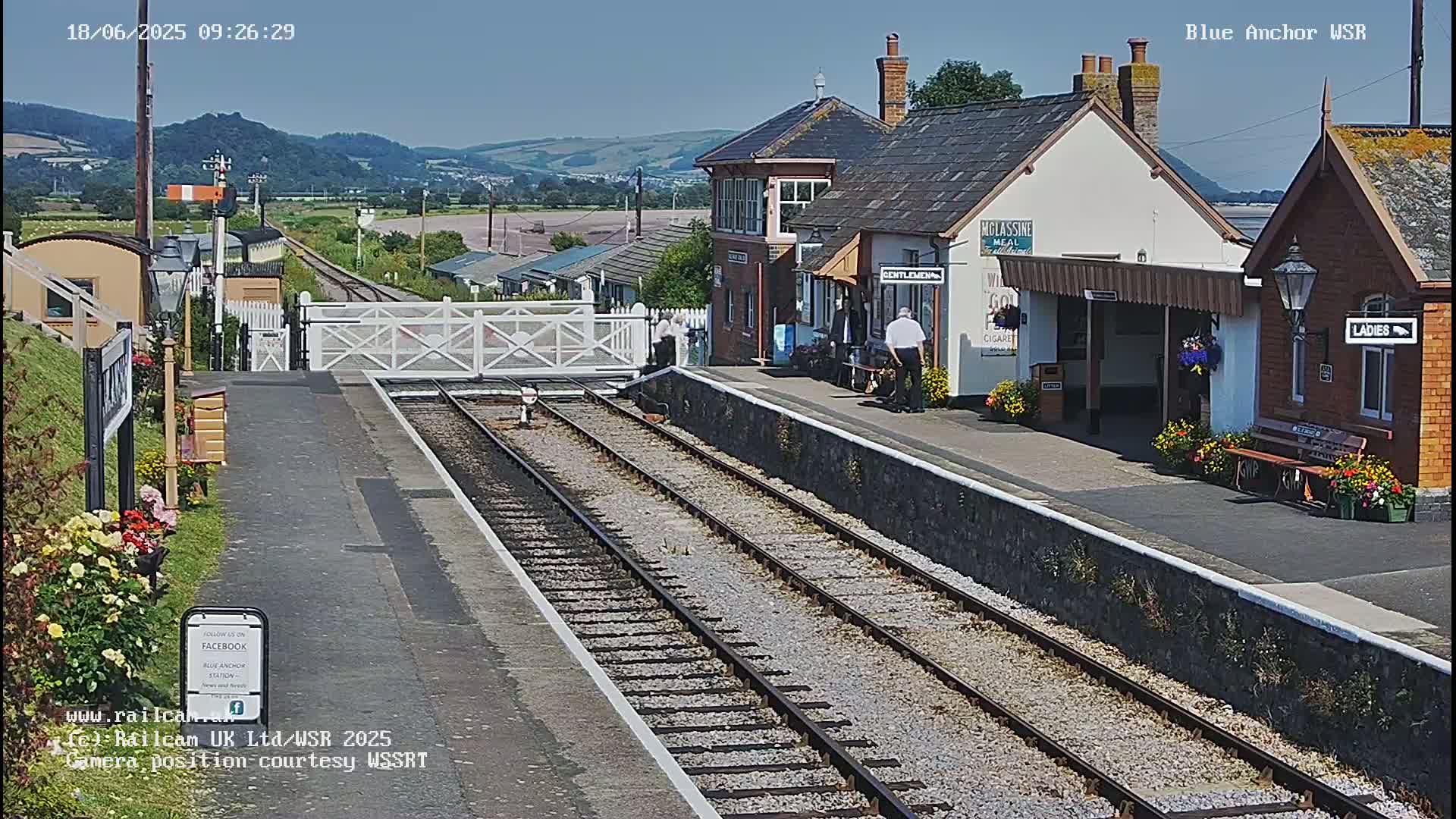 A train station with two sets of tracks and several buildings, including a signal box, under a clear, sunny sky with distant hills.