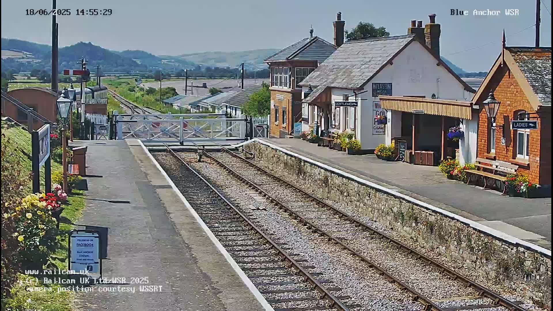 A sunny view of a train station with two platform buildings, a level crossing, and train tracks stretching into the distance, backed by rolling hills.