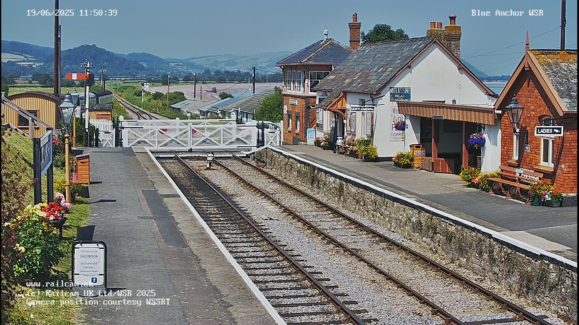 A sunny view of a train station with a level crossing gate, railway tracks, and station buildings with flower boxes.