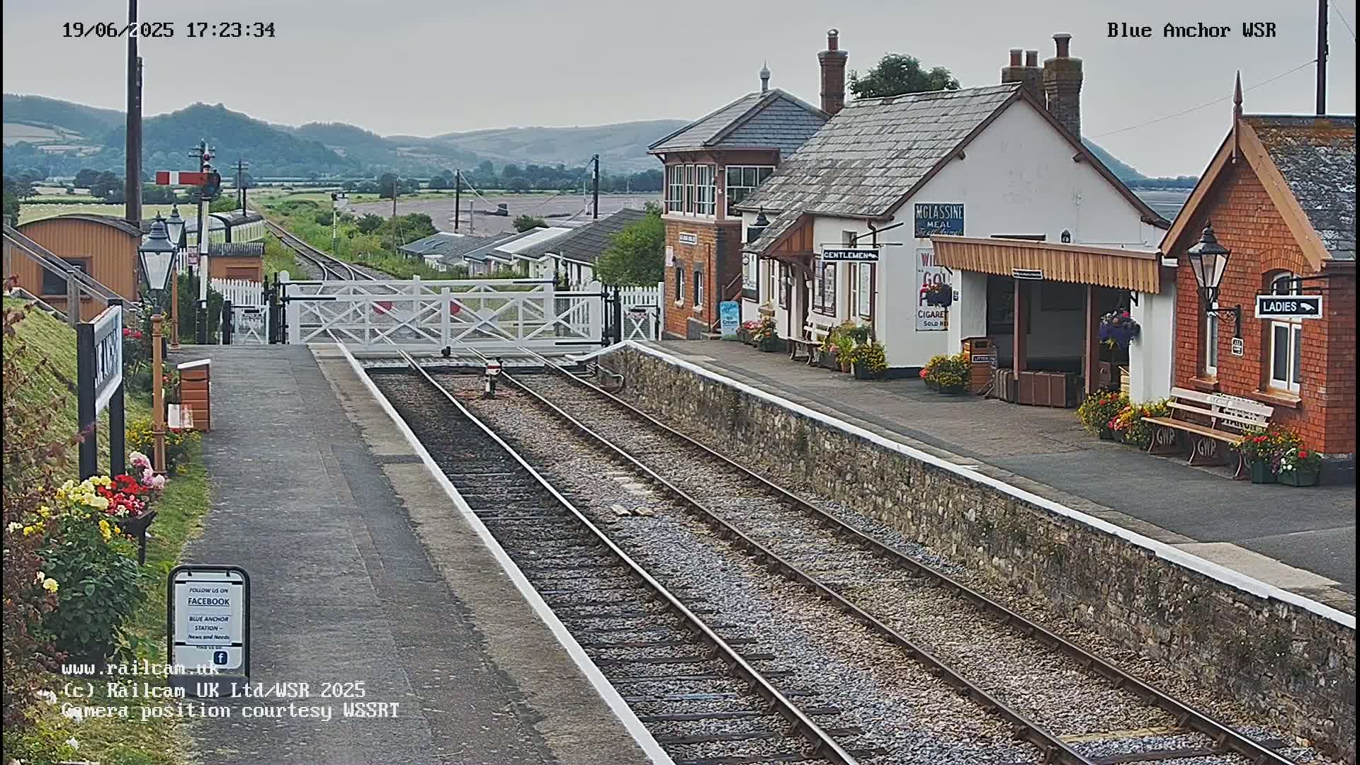 A train station with a signal box and platform sits under an overcast sky, with hills visible in the background.