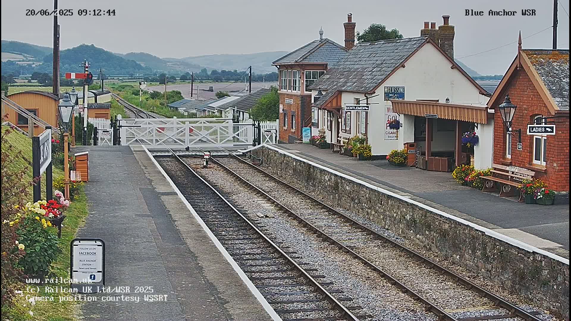 A train station with a brick building and waiting shelters sits under an overcast sky next to a railway track.