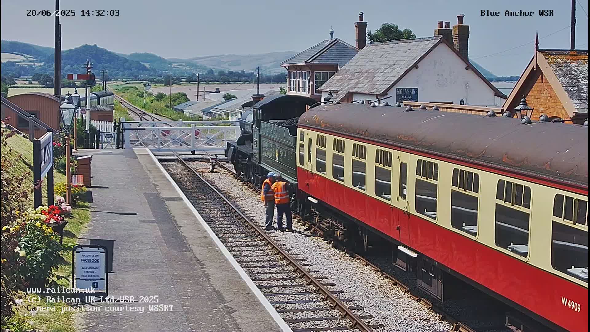 A red and cream passenger car attached to a dark green steam engine sits at a train station on a sunny day, with rolling hills visible in the background.