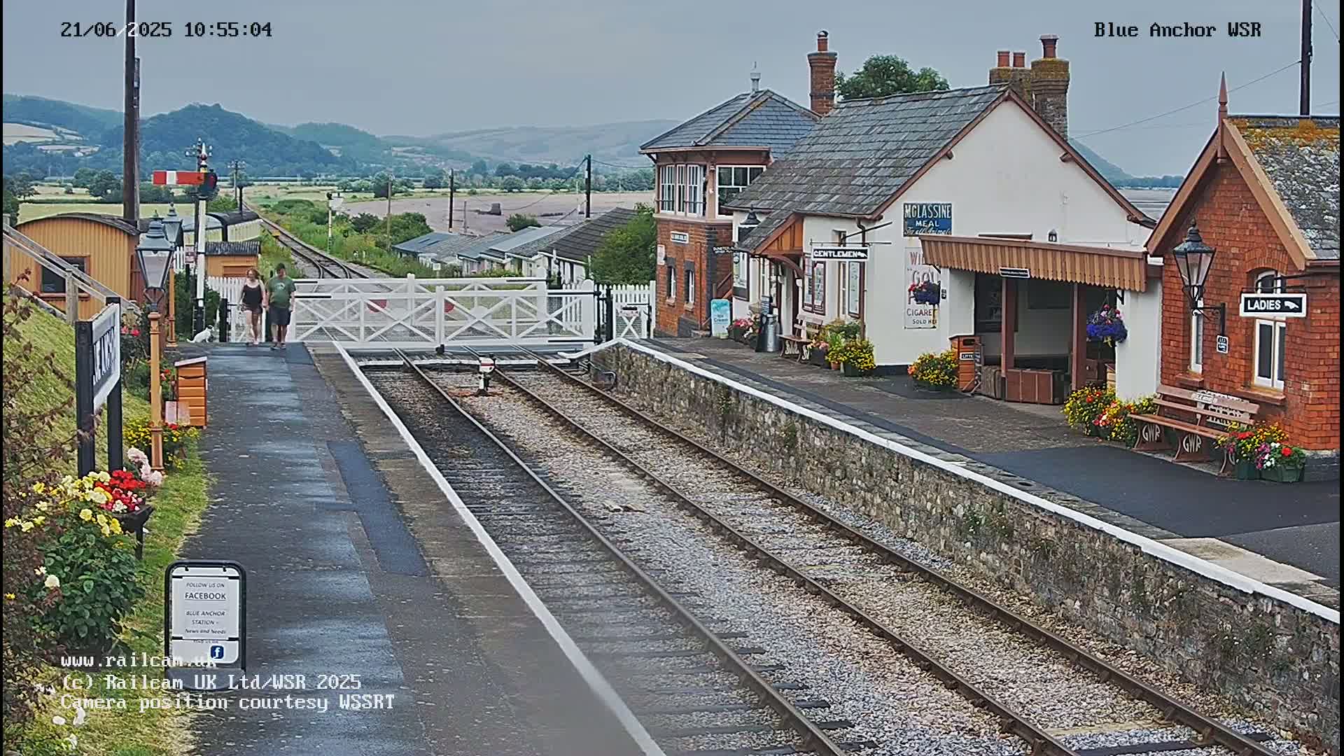 A train station with two buildings and a platform is seen on an overcast day, with train tracks extending into the distance.
