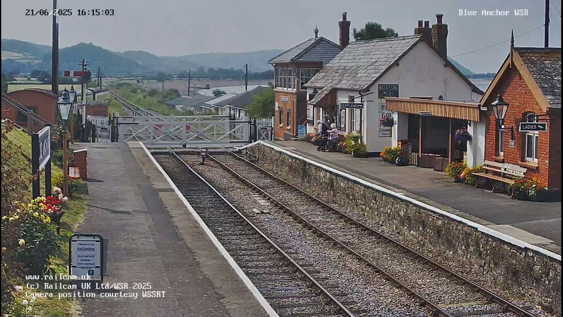 A train station platform with two sets of tracks, railway buildings, and a few people, under a clear, sunny sky and distant hills.