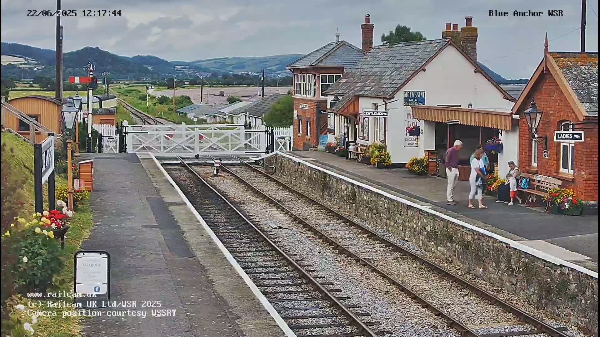 A train station with several buildings, a few people standing on the platform, and a railway track leading to a level crossing under a partly cloudy sky.