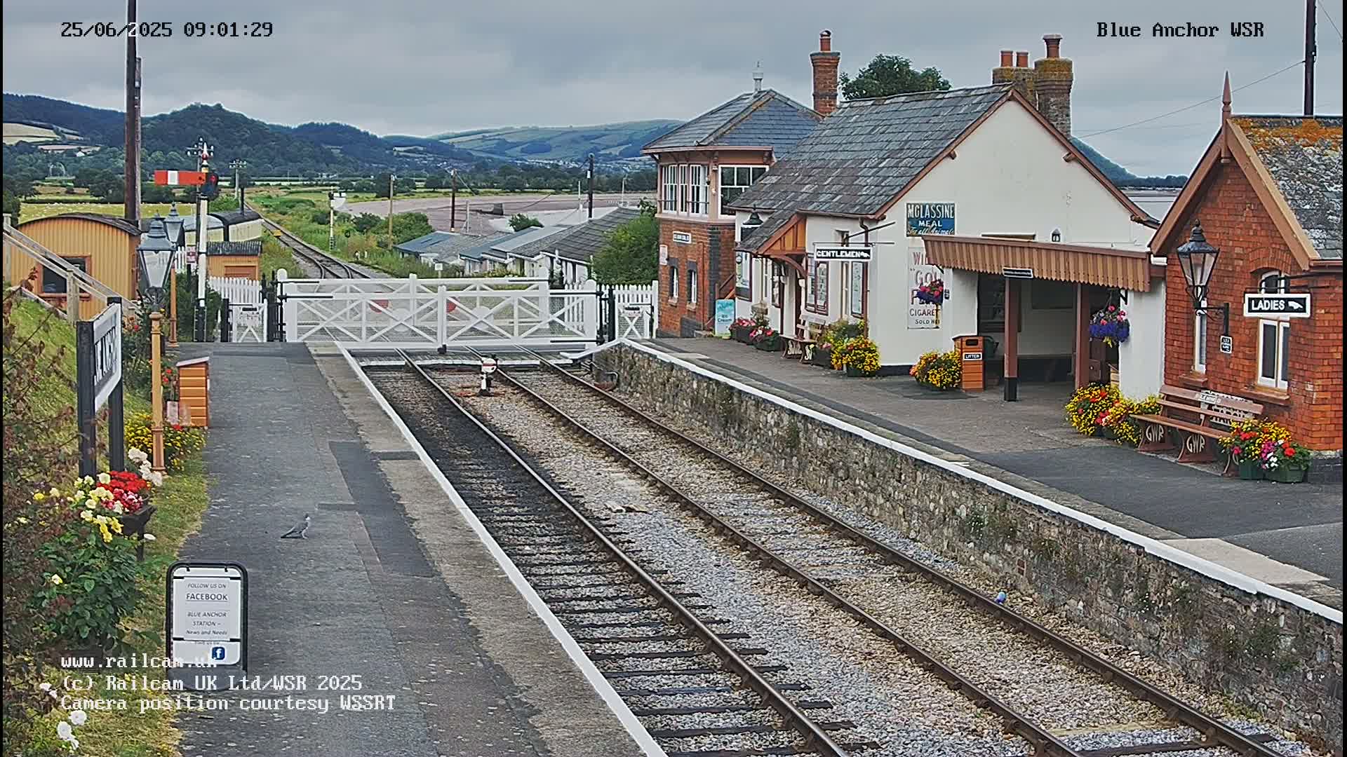 A train station with a signal box and waiting shelters is viewed from the tracks on an overcast day.