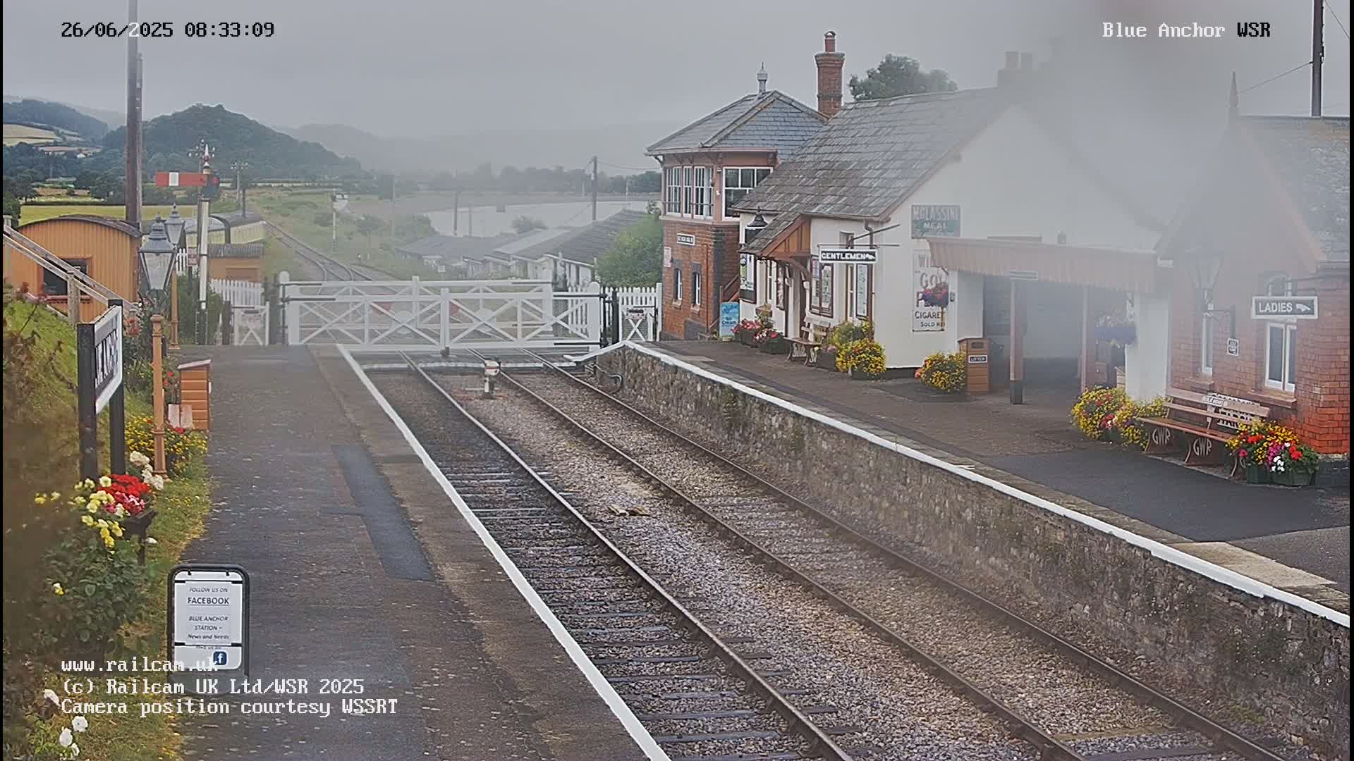 A foggy view of a train station with two train tracks, a signal box, and a platform lined with flower boxes.
