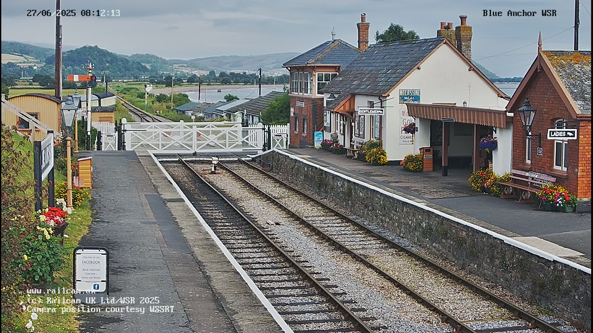 A train station with a low stone platform and railway tracks extends into the distance, under an overcast sky, toward a body of water and hills.