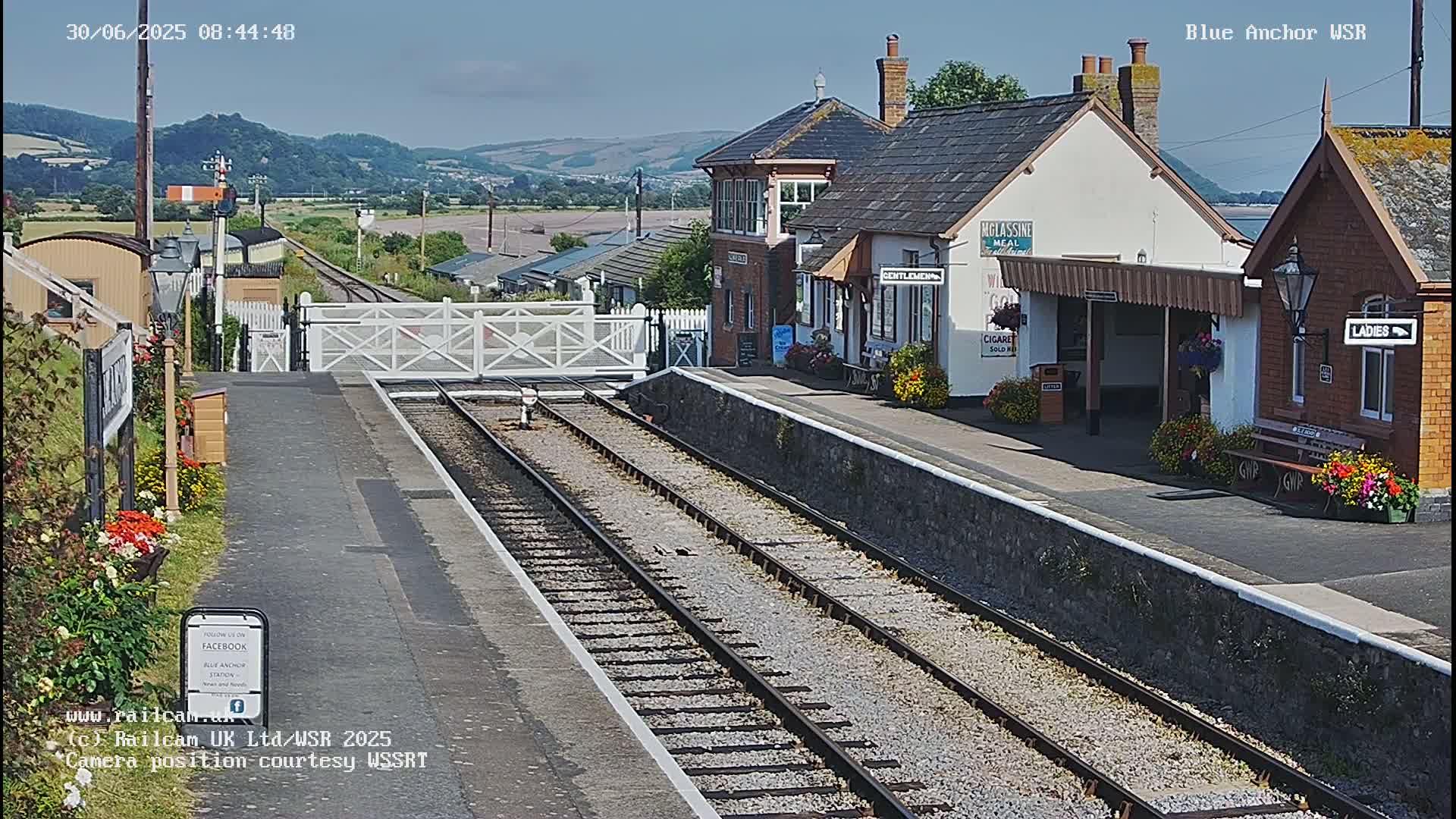 A train station with a signal gate and several buildings, situated on a sunny day with hills visible in the background.
