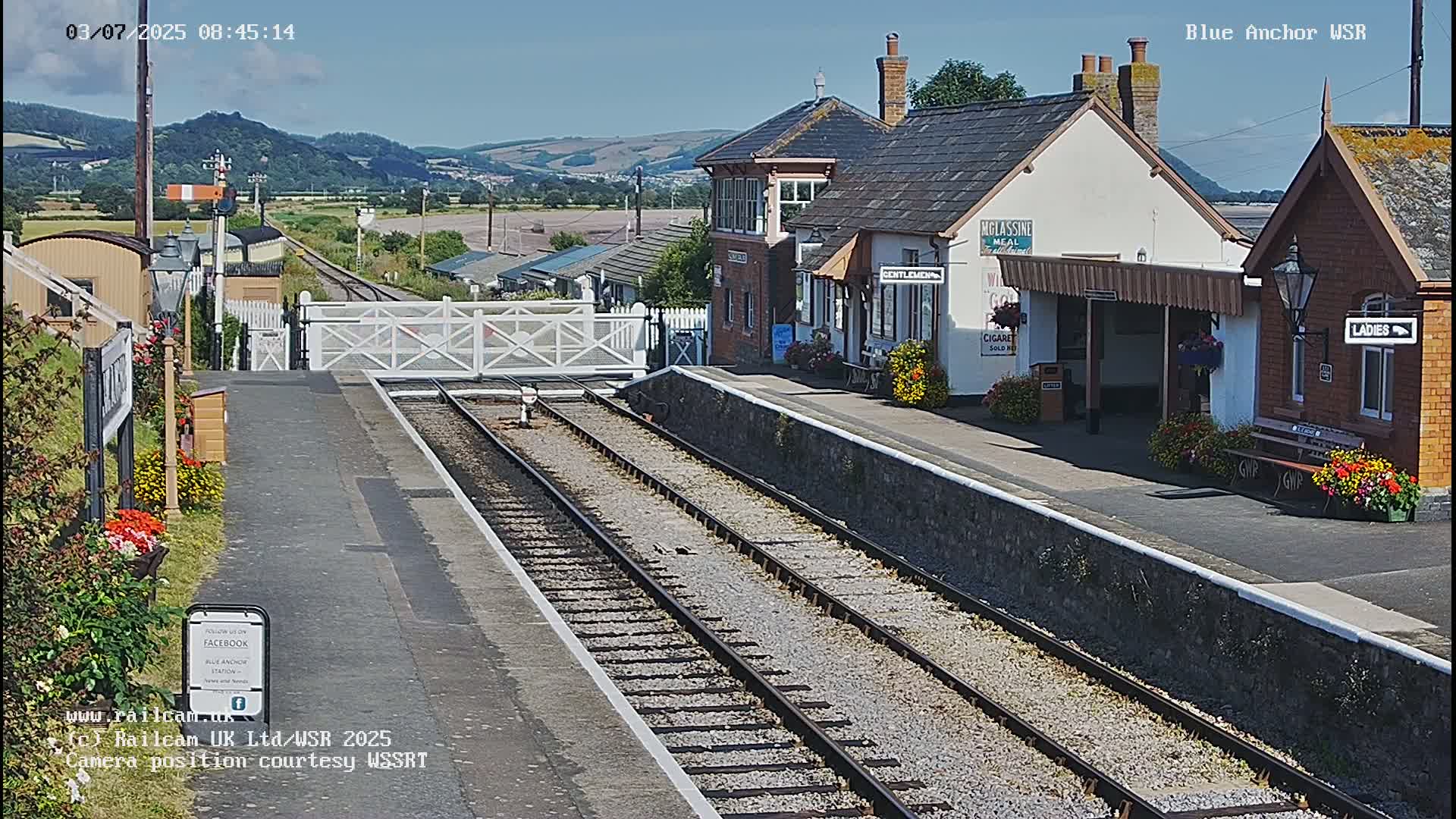 A sunny view of a train station with two tracks leading to a closed gate, featuring a signal box and small station buildings, surrounded by flowerbeds.