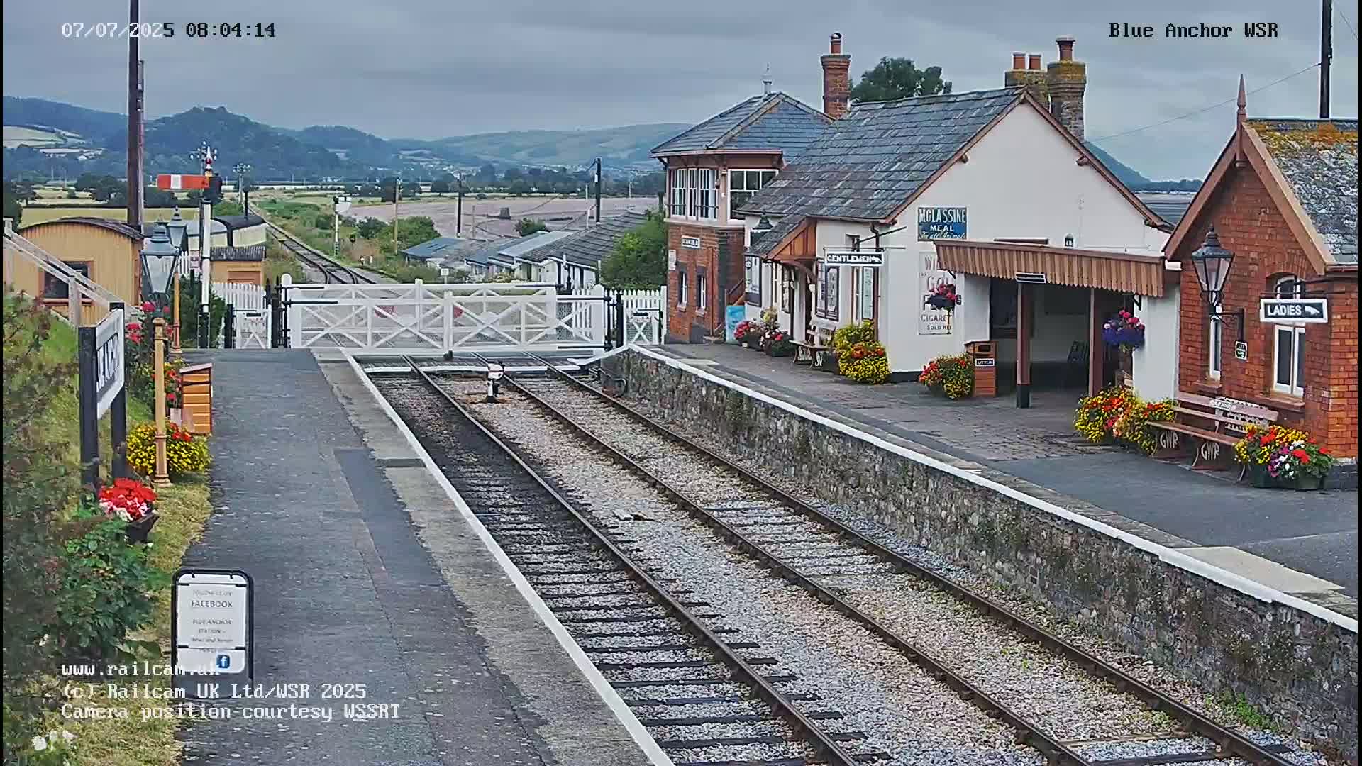 A train station with two sets of tracks, a platform, and station buildings, under an overcast sky.