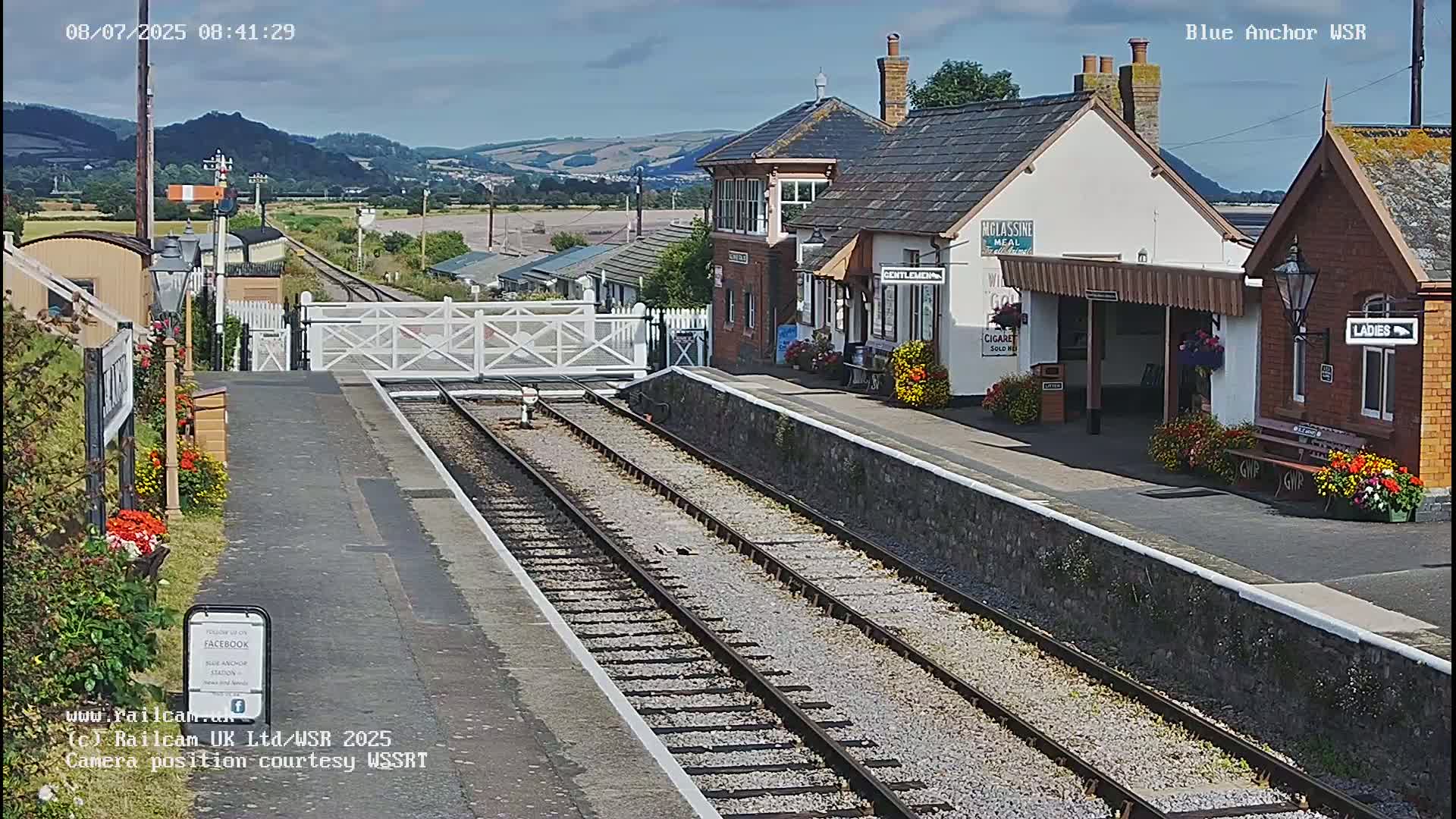 A train station with a small building and platform, under sunny skies, is situated in front of rolling hills.